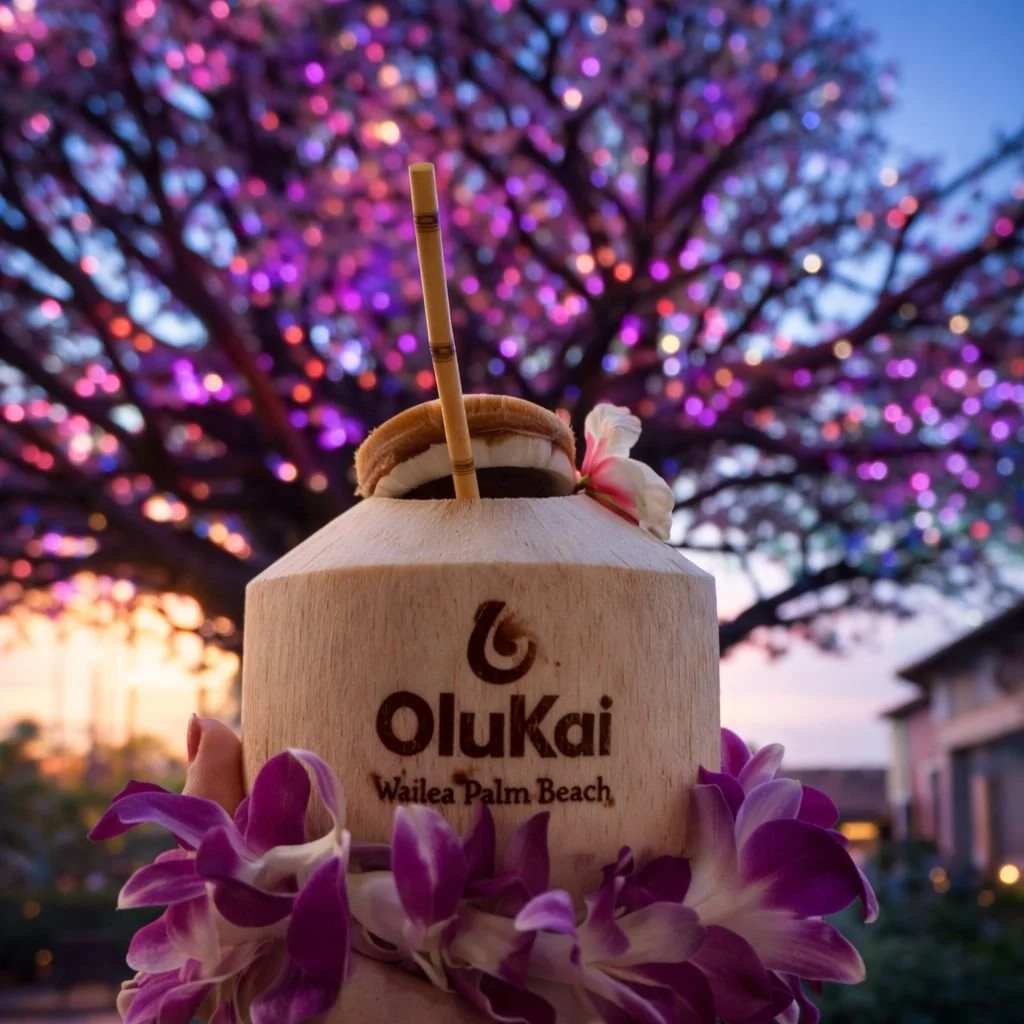 A coconut drink with a straw and flower, branded 'OluKai Wailea Palm Beach', held up in front of a blooming purple and pink tree during sunset.