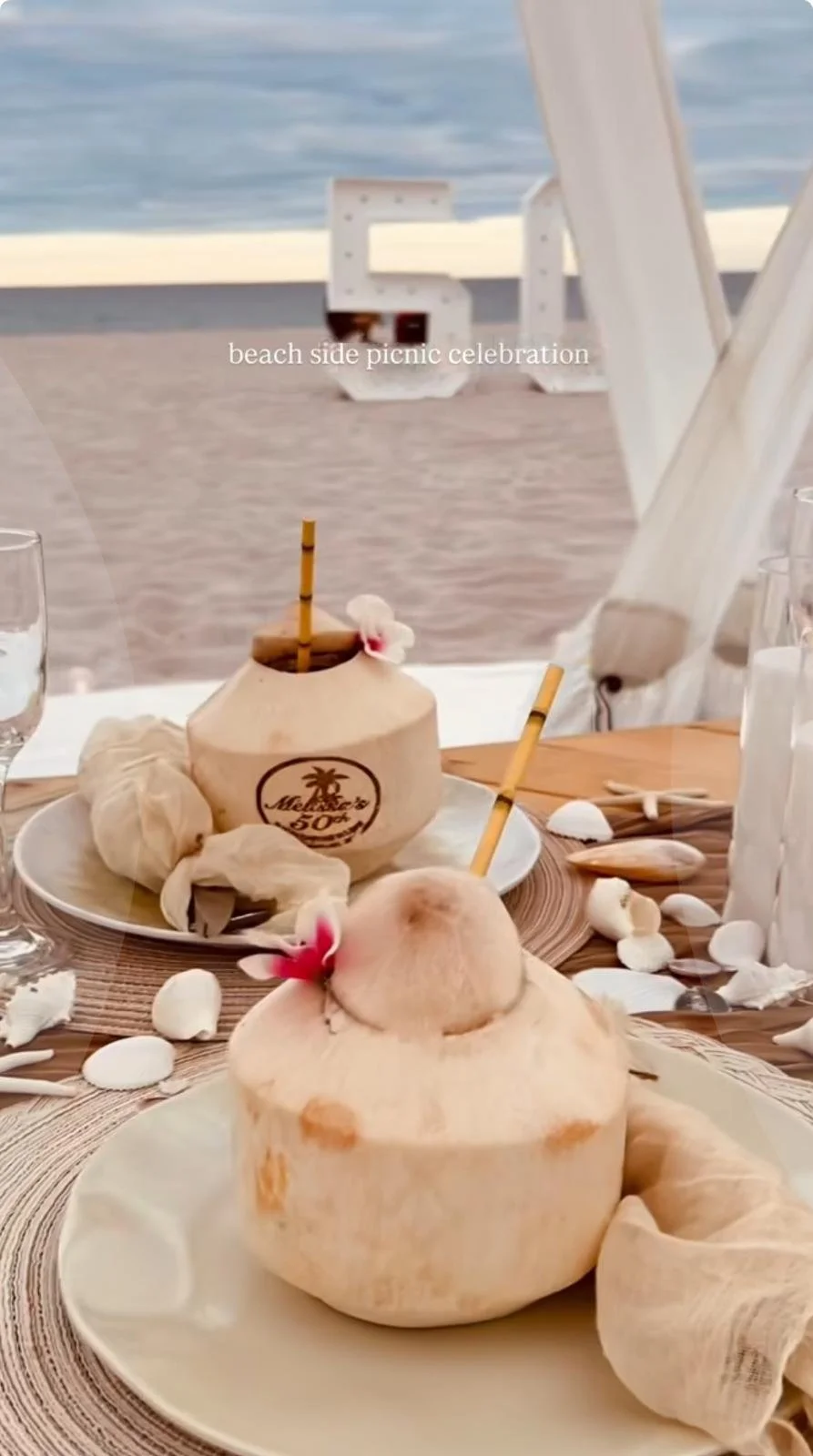 Coconut drinks and seashells on a beachside table with ocean in the background, decorated for a beach side picnic celebration.
