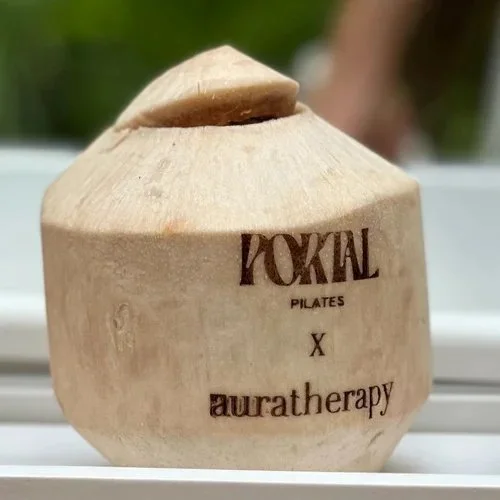 A wooden coconut with the words "POKAL PILATES X auratherapy" burned into it, sitting outdoors on a white surface, with a blurred background of green foliage.