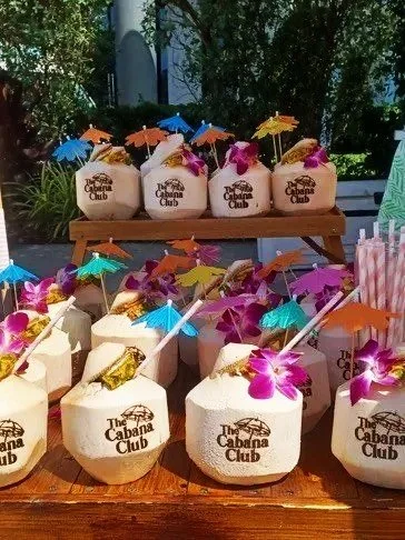 Coconuts with drink umbrellas and orchids on a wooden table outdoors, branded with 'The Cabanas Club' logo.