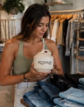 Woman shopping in a clothing store holding a coconut drink with a straw labeled 'KOPARI'.