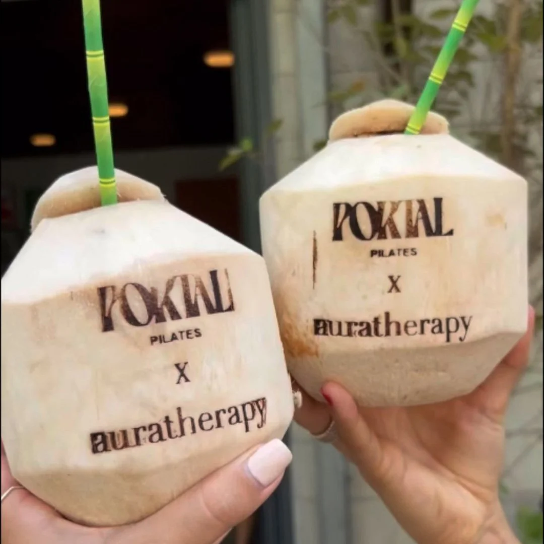 Two hands holding coconut drinks with straws, branded with 'TOKAI PILATES X auratherapy,' in an outdoor setting.