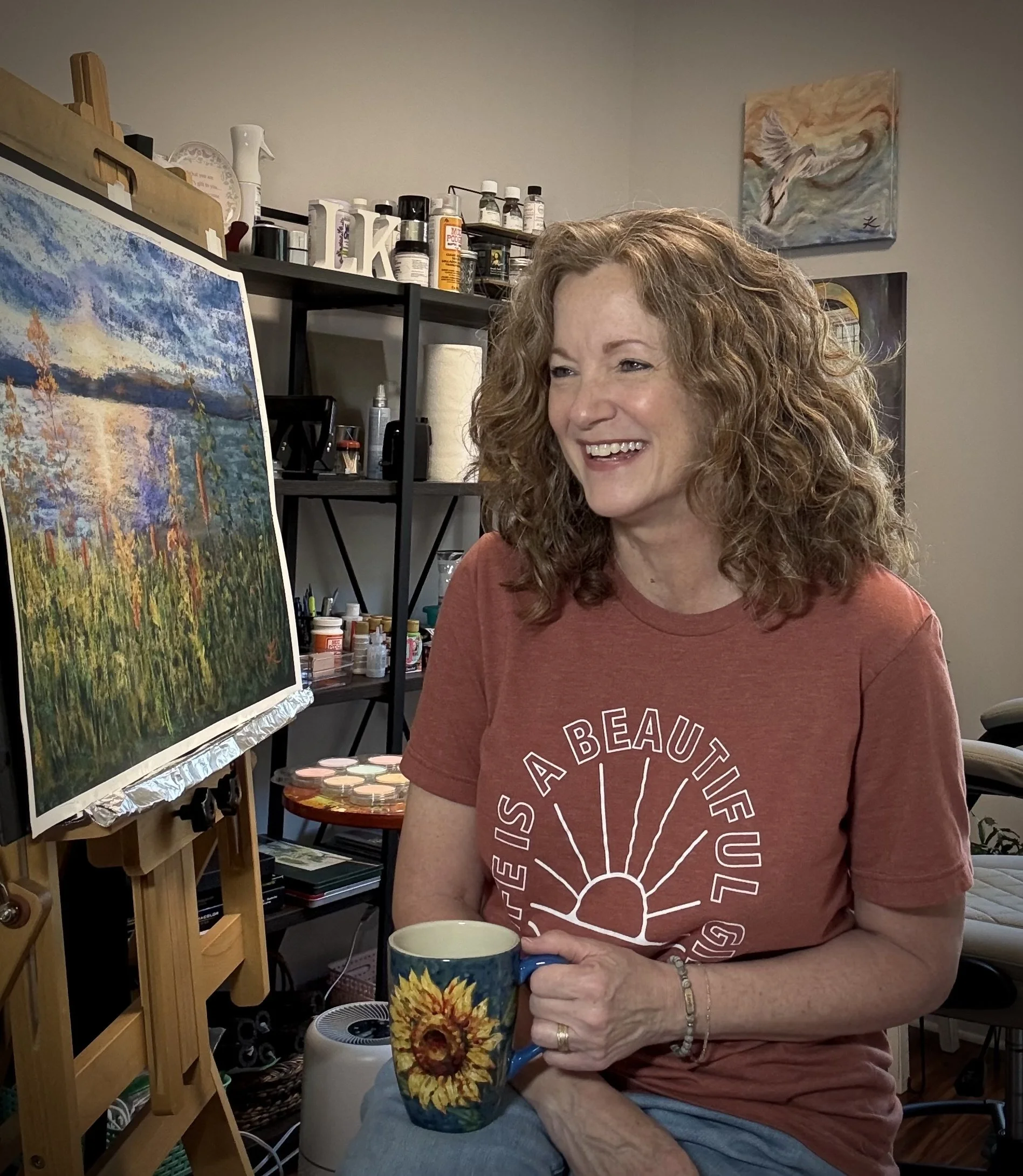 Auburn-haired woman sitting in front of her art easel, smiling and holding a cup of coffee