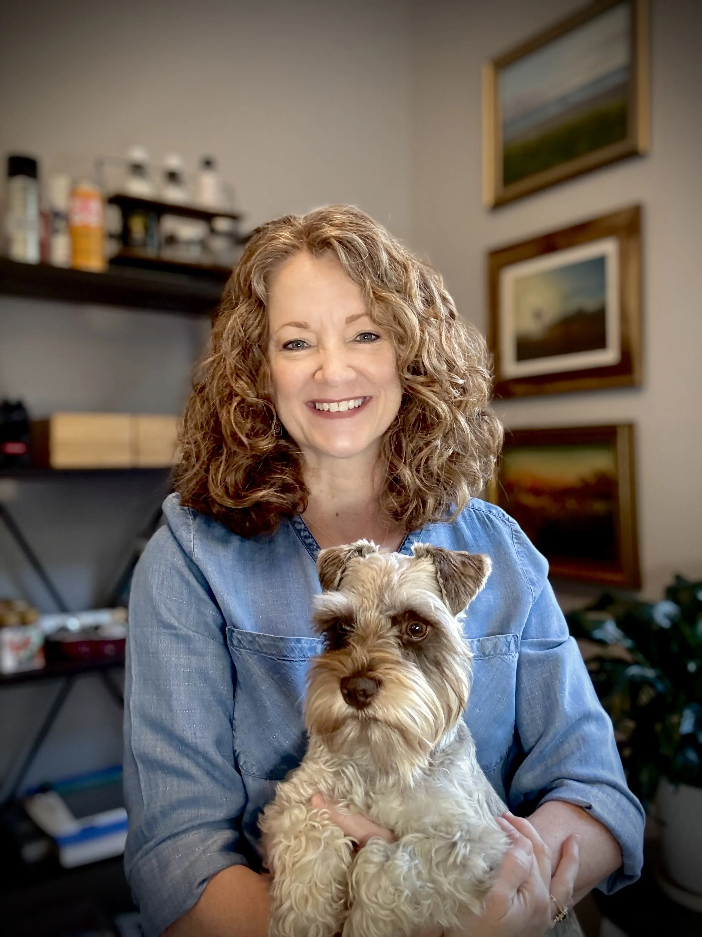 A woman is sitting in her art room, holding her miniature schnauzer in her lap.