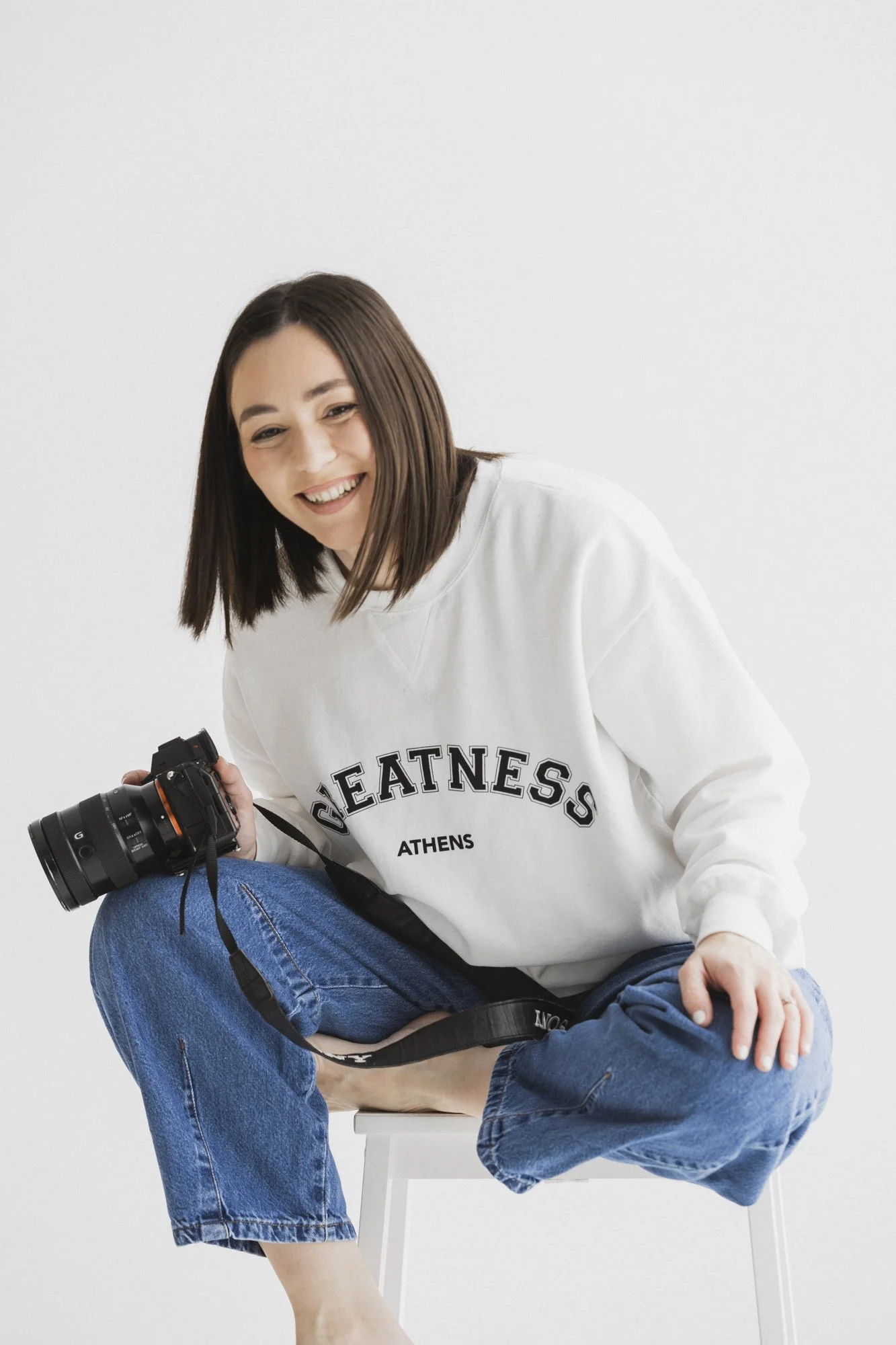 Smiling person in a white "Greatness Athens" sweatshirt holding a camera, sitting on a stool against a white background.