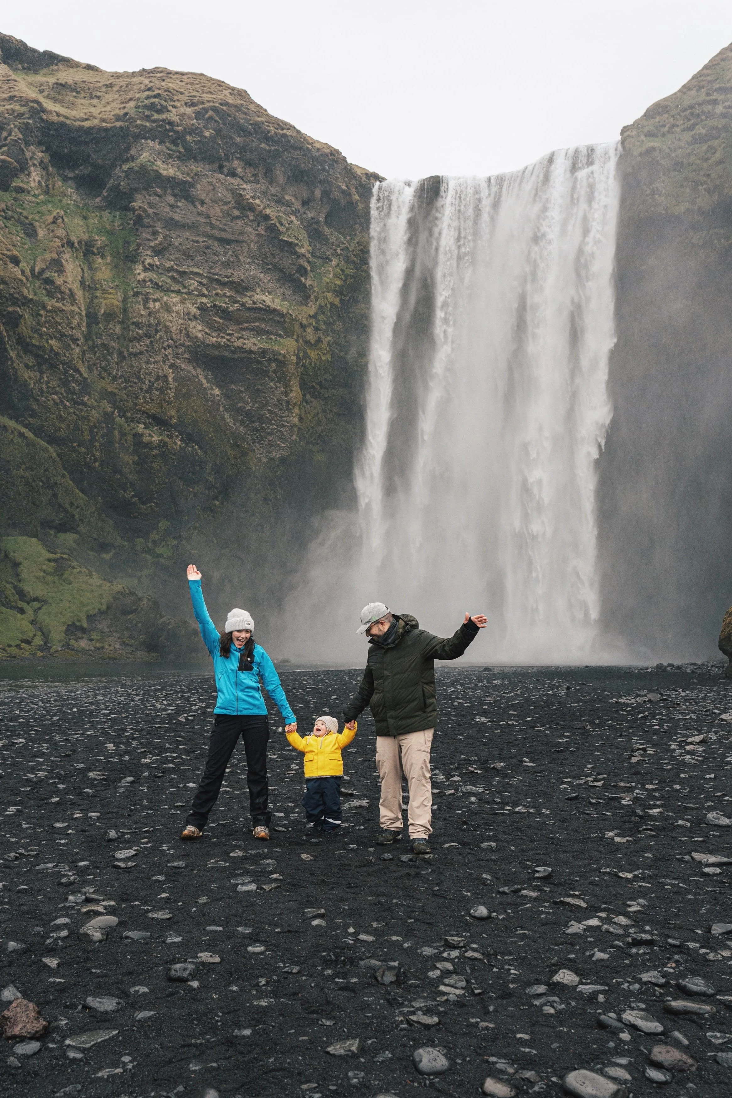 Family with a child posing in front of a large waterfall on a rocky black sand landscape.