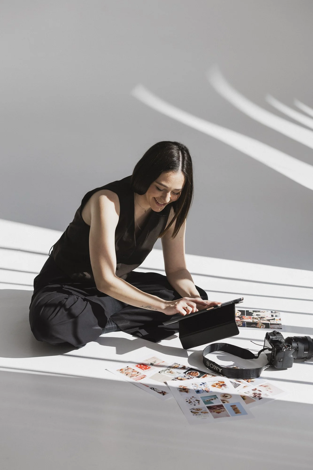 Woman sitting on the floor, using a tablet. Assorted photos and a Sony camera are placed nearby. Bright lighting with shadow patterns cast across the scene.