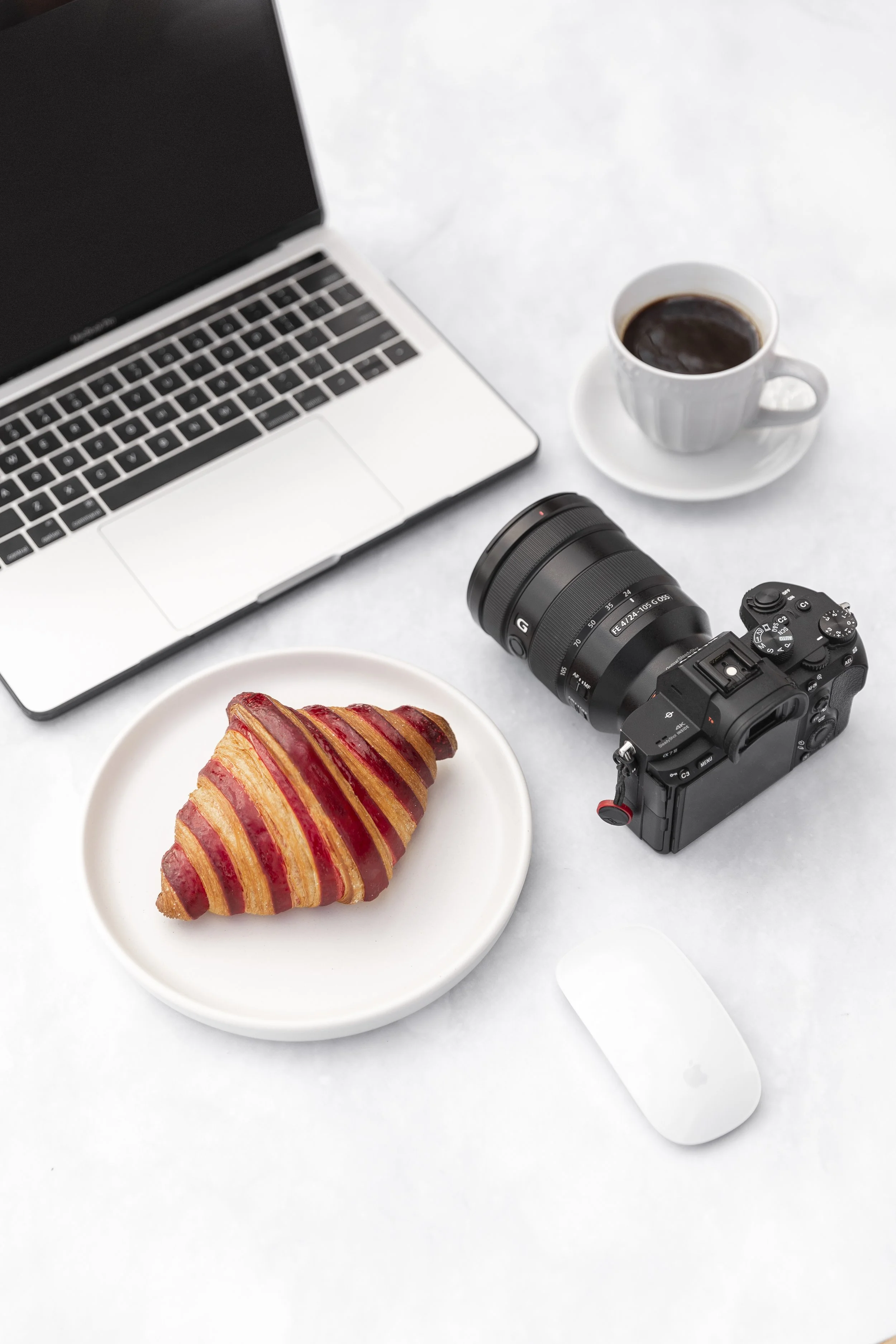 A laptop, a croissant on a plate, a camera with lens, a cup of coffee, and a wireless mouse on a white surface.