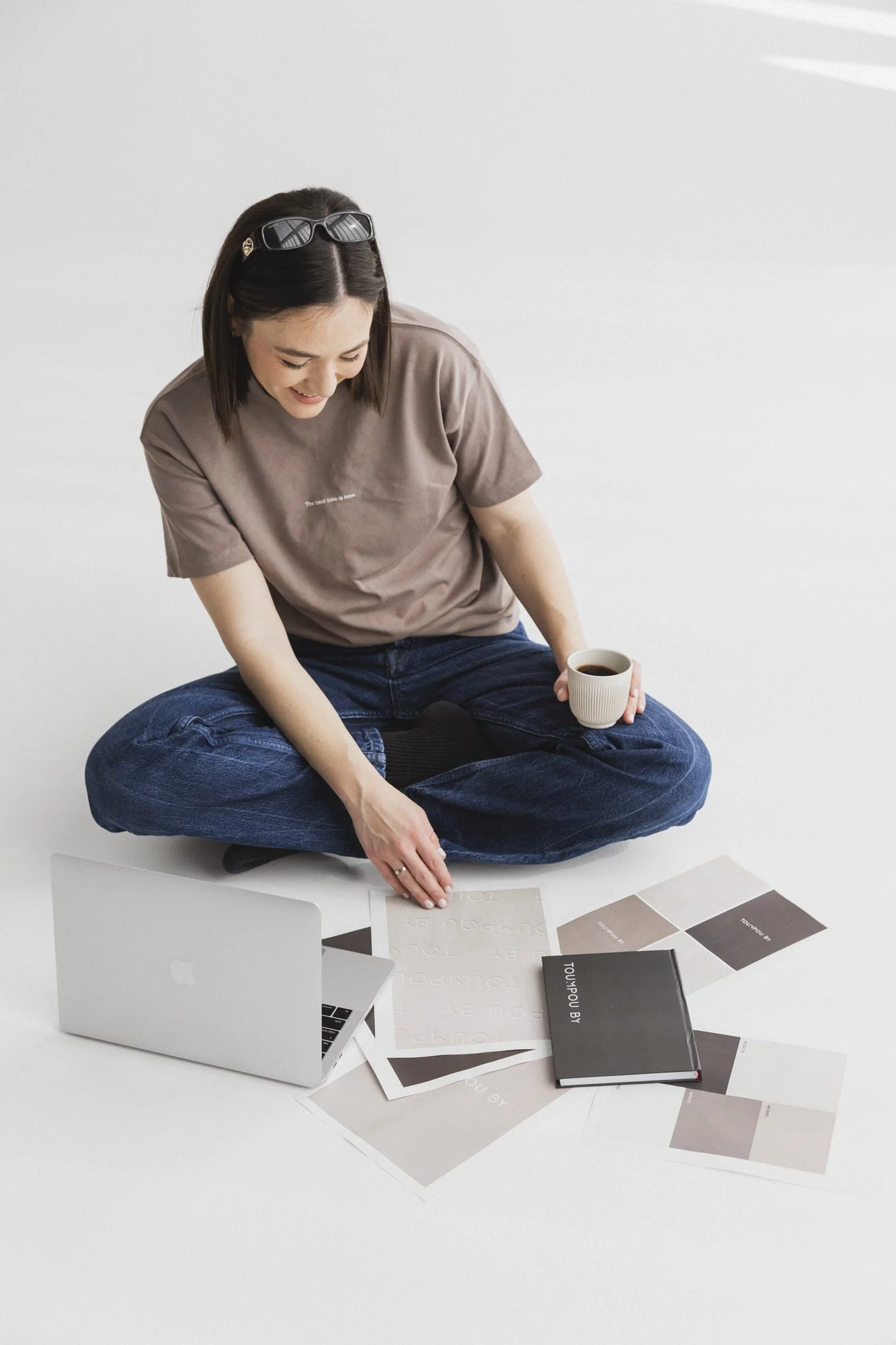A person sitting cross-legged on the floor with a laptop, holding a coffee cup, surrounded by design samples and a notebook.