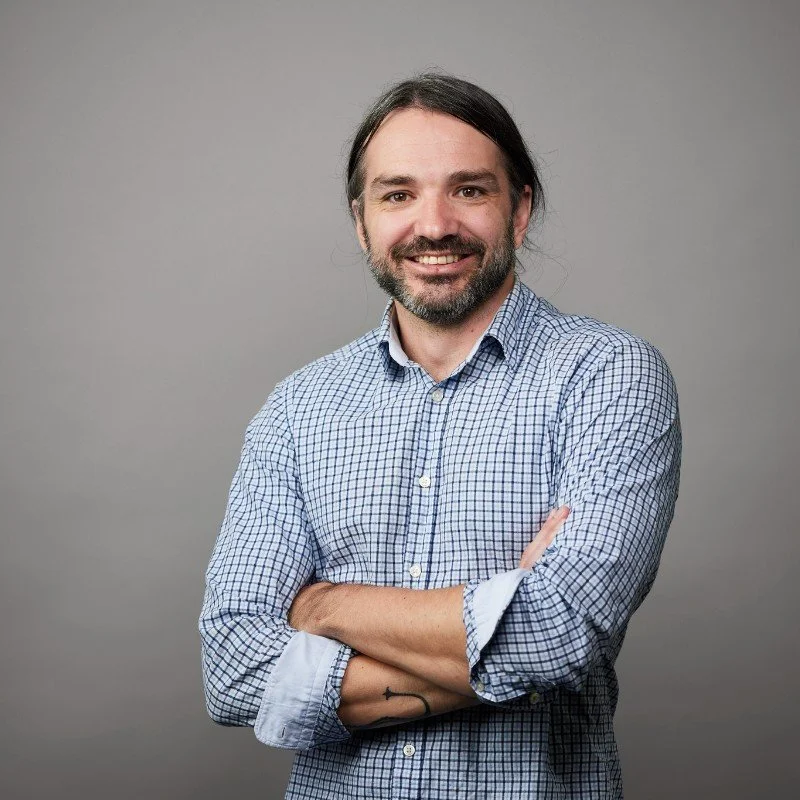 A photo of Ray, a smiling man with long dark hair, beard, and mustache wearing a blue checkered button-up shirt with rolled-up sleeves standing against a plain gray background.