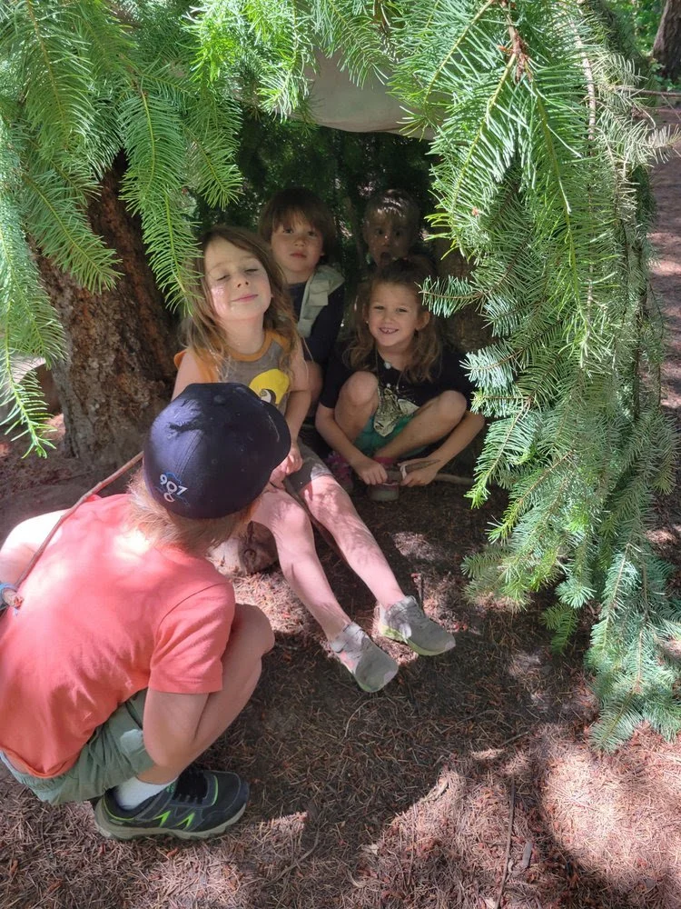 Group of children playing in a small makeshift fort or hiding spot under a tree, with a boy crouching outside and four girls sitting inside.