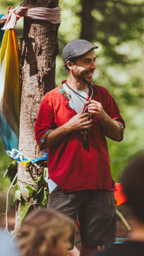 Man smiling outdoors, wearing a gray cap and red shirt, holding prayer beads near a tree.