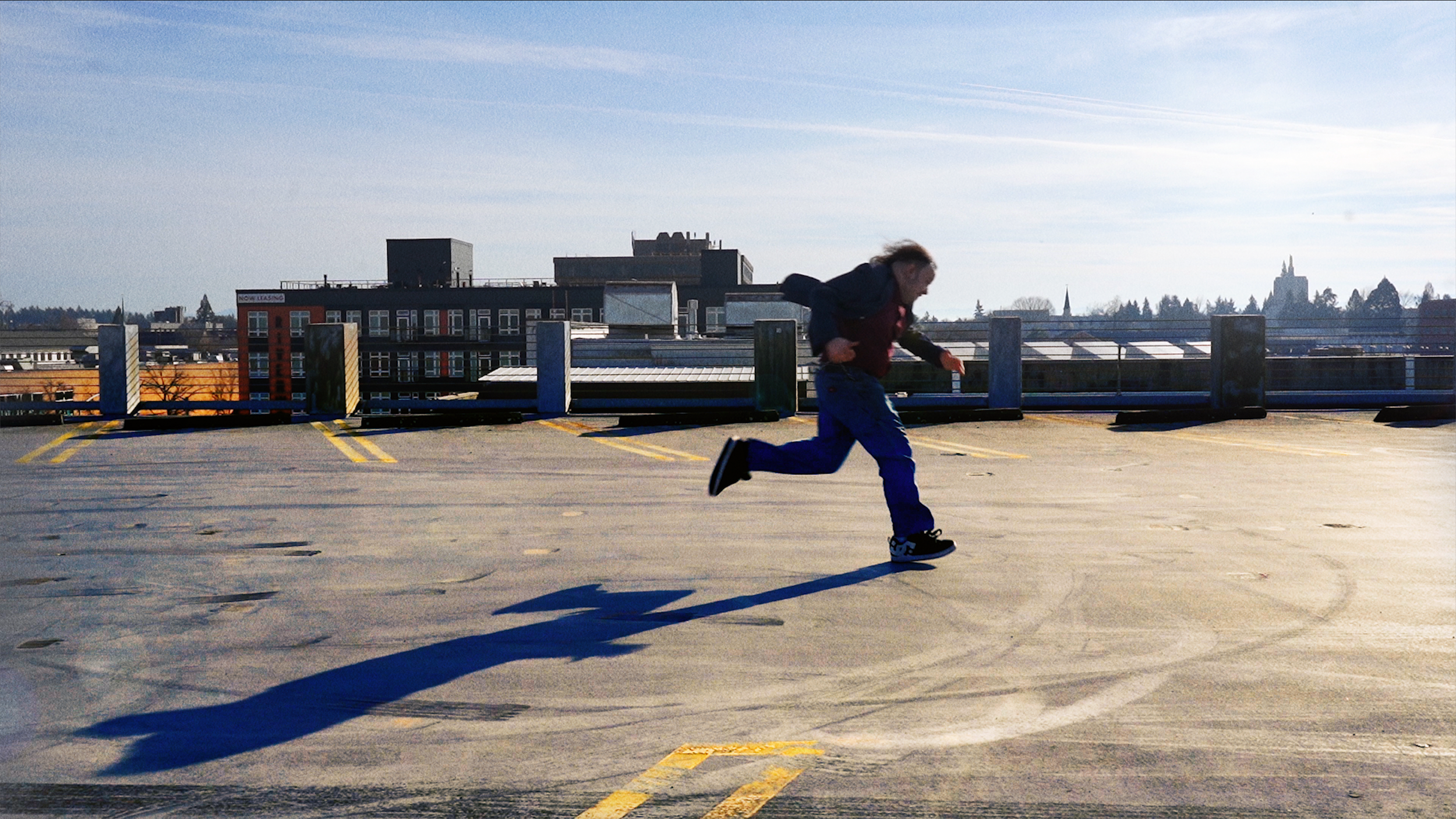 A person running on an empty rooftop parking lot during the daytime, with buildings and a clear sky in the background.