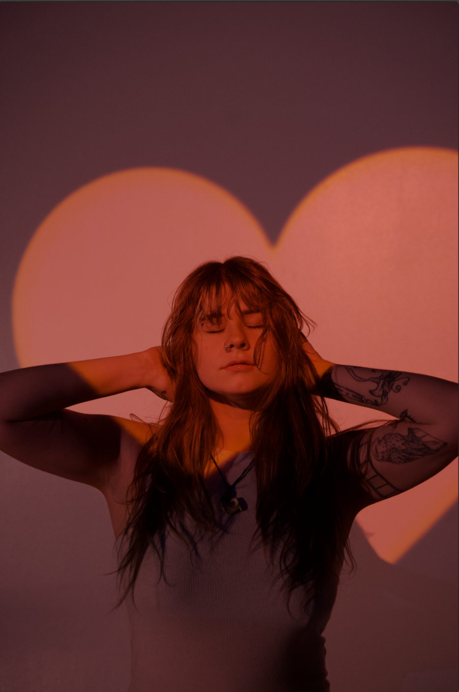 A woman with red hair and tattoos on her arms, eyes closed, with hands behind her head against a background of a heart shape projected in pink and orange light.