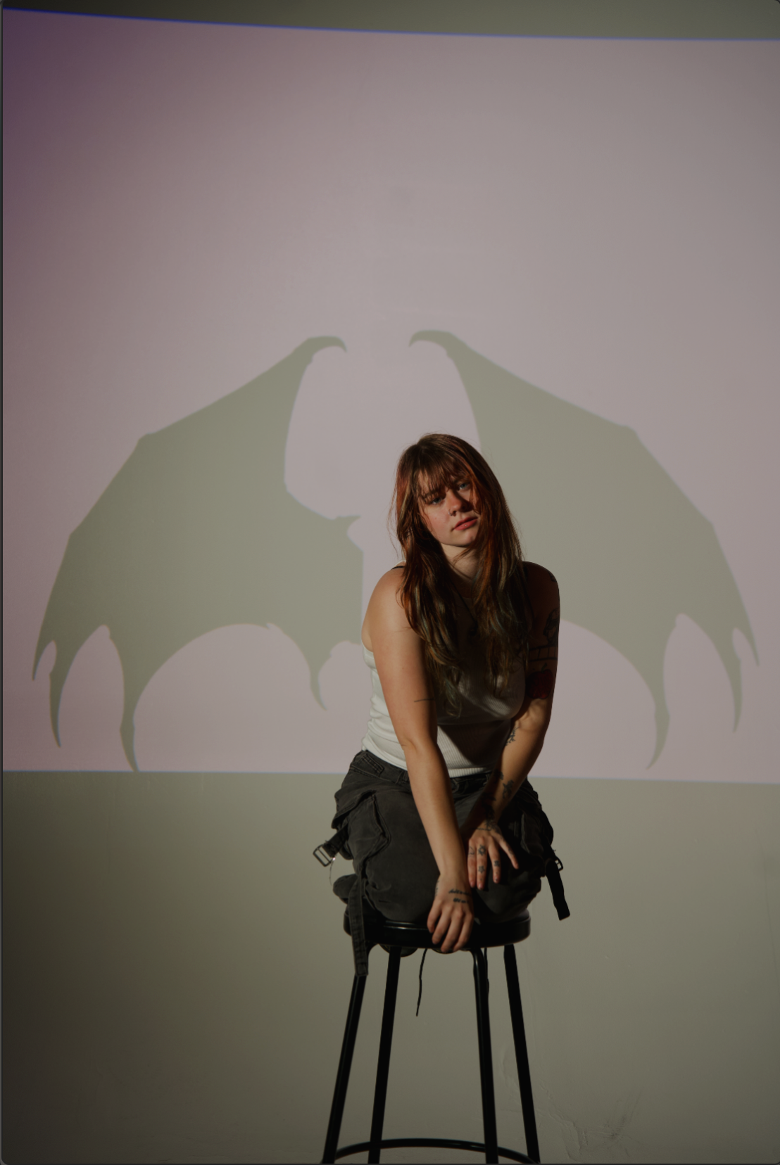 A young woman with long hair and tattoos, wearing a white tank top and black cargo pants, sitting on a stool with her knees bent and hands resting on her knees. Behind her, a shadow of demon wings is projected on the wall.