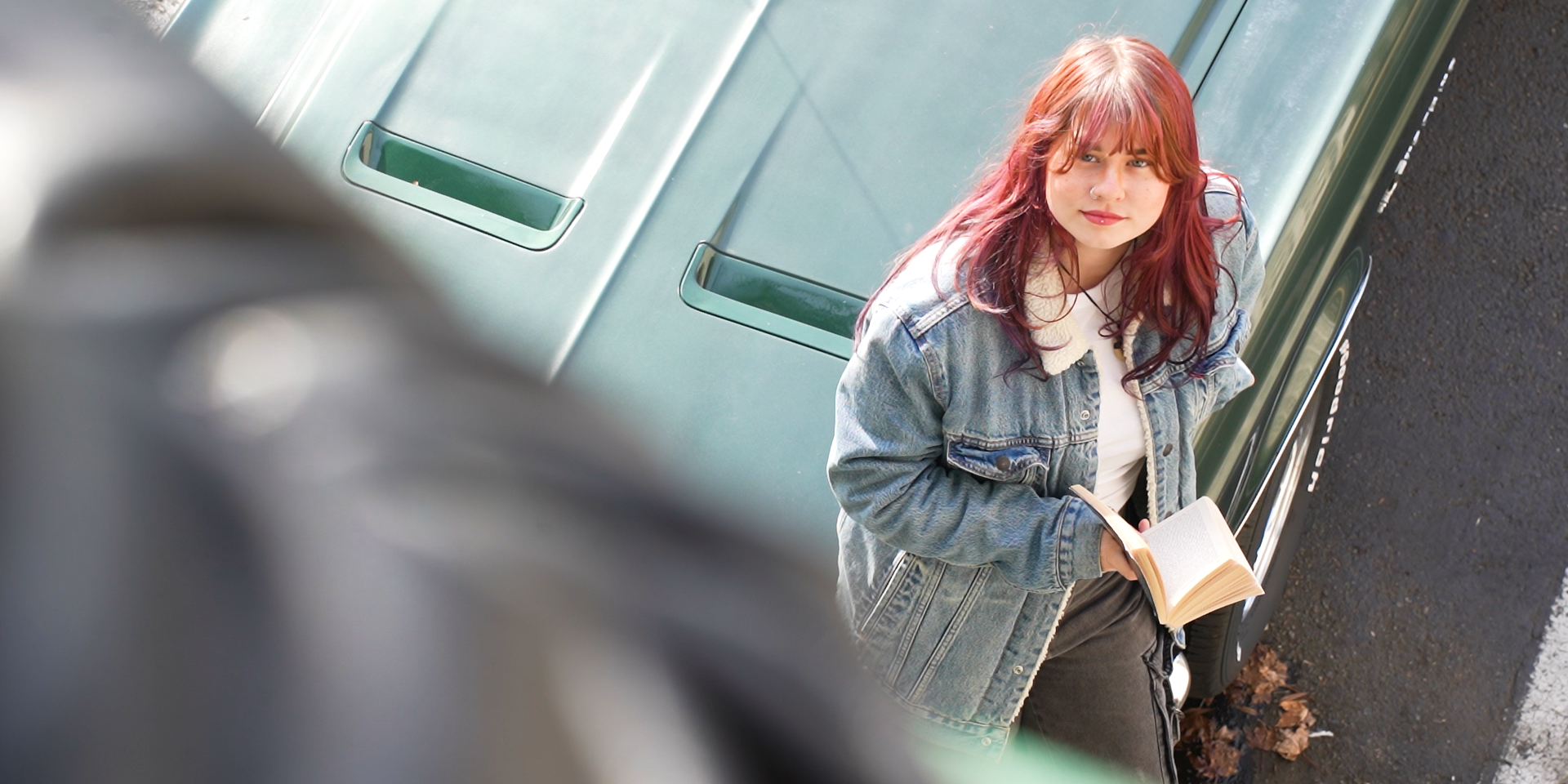 A young woman with red hair holds an open book and looks up toward the camera, standing outside next to a dark green car parked on a paved road.