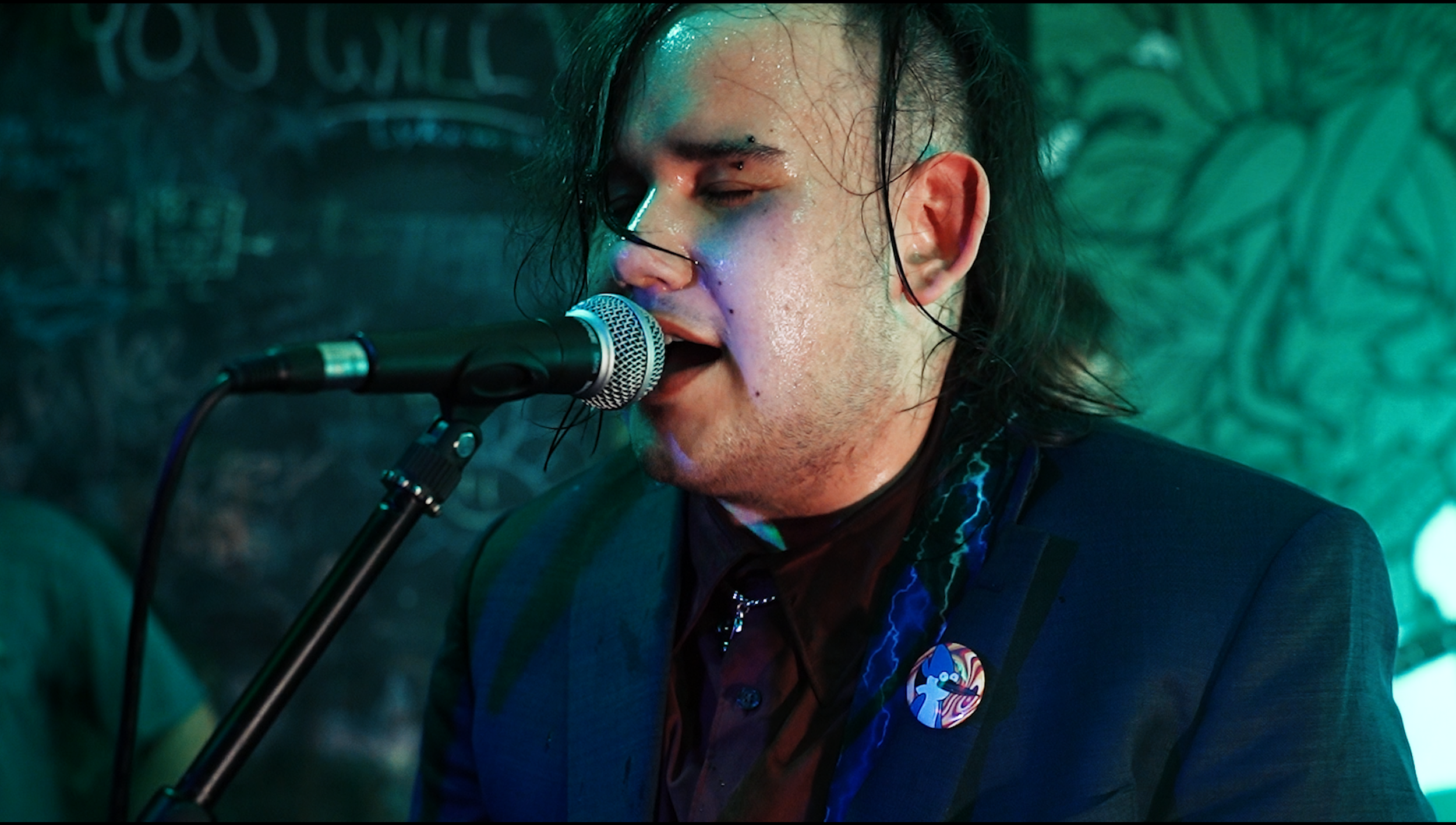 A man singing into a microphone at a performance with a background of green and blue textured artwork.