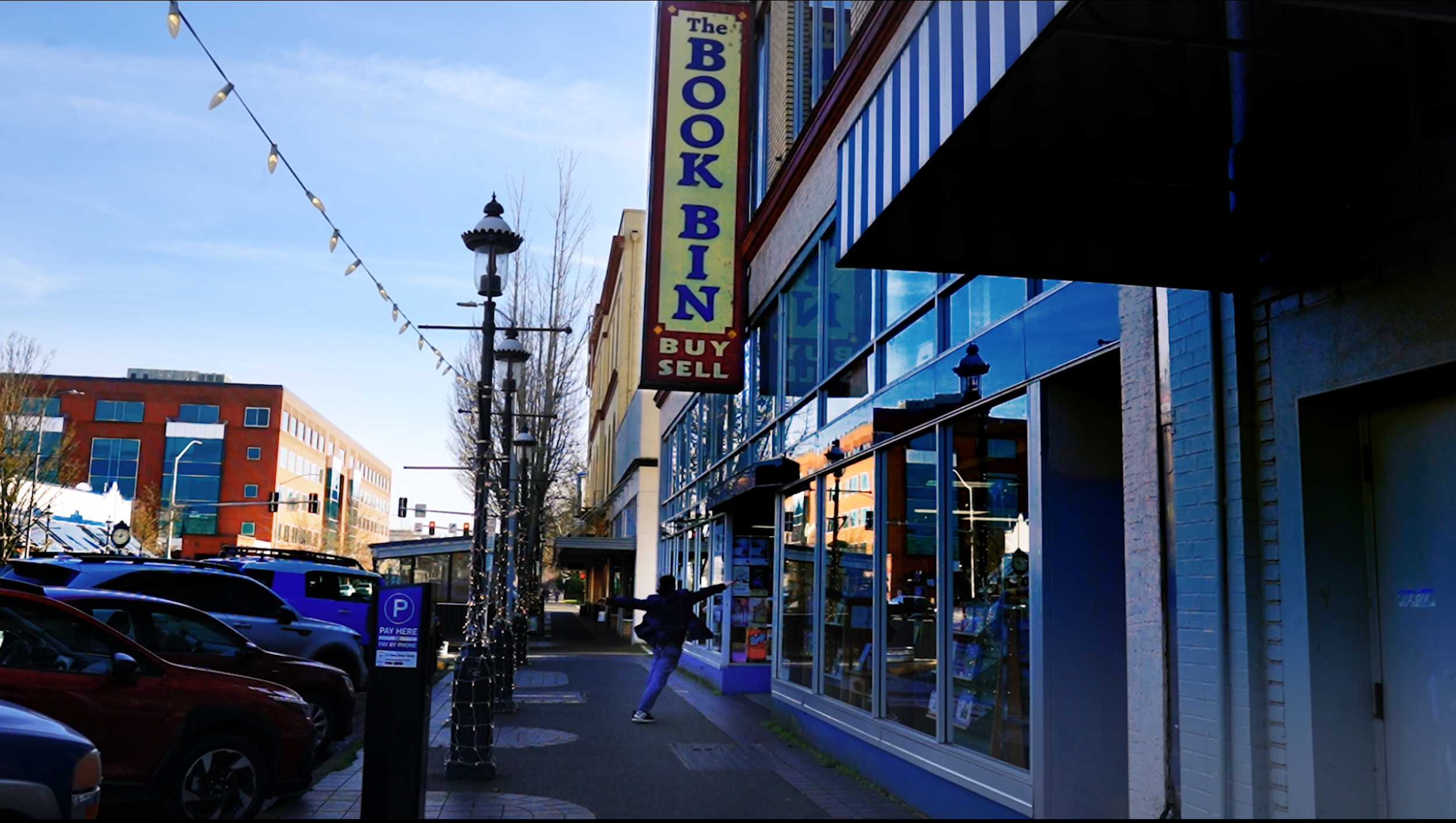 Street view with a sign for a bookstore that sells and buys books, parked cars, and a person reaching out towards the store.
