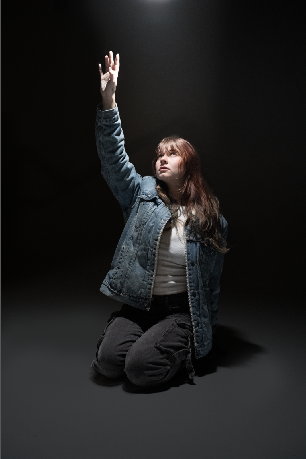 A young woman with red hair, wearing a denim jacket and dark pants, kneeling on the ground and reaching upward with her right hand in a dark, minimal setting.