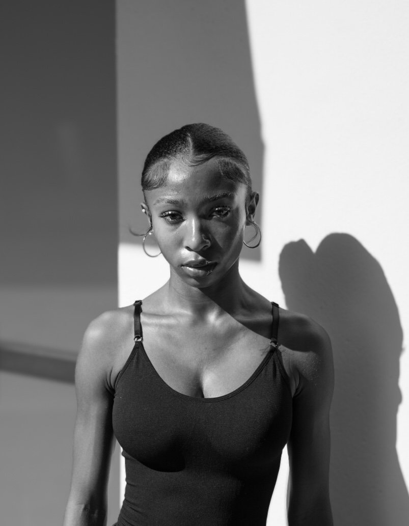Black and white portrait of a woman with hoop earrings, wearing a tank top, standing against a wall with a shadow.