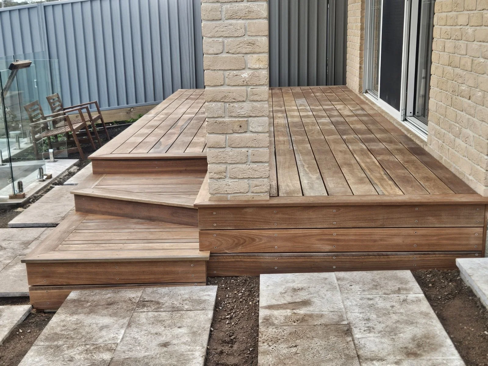 Wooden deck with steps and pavers, adjacent to a brick wall and glass railing.