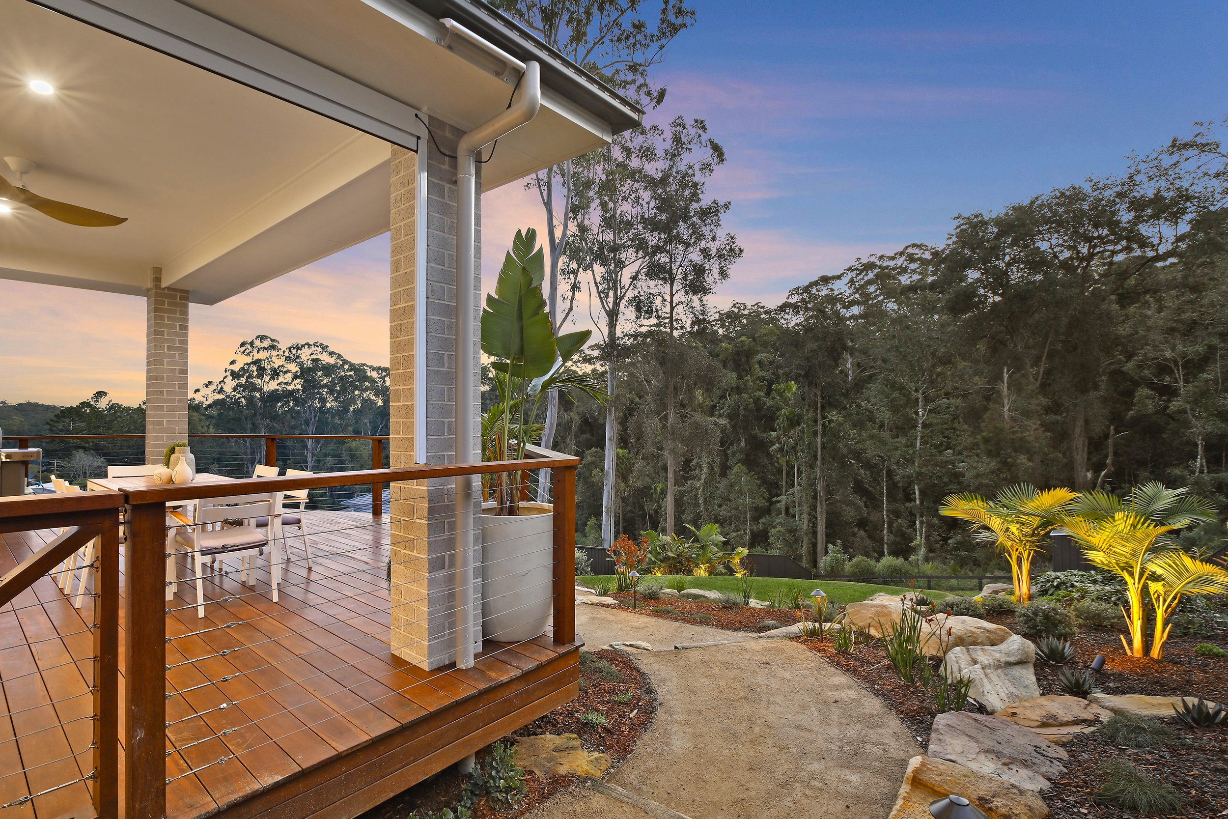 Outdoor wooden deck with dining area and chairs, surrounded by lush greenery and trees, with a view of a landscaped garden path and ambient evening lighting.