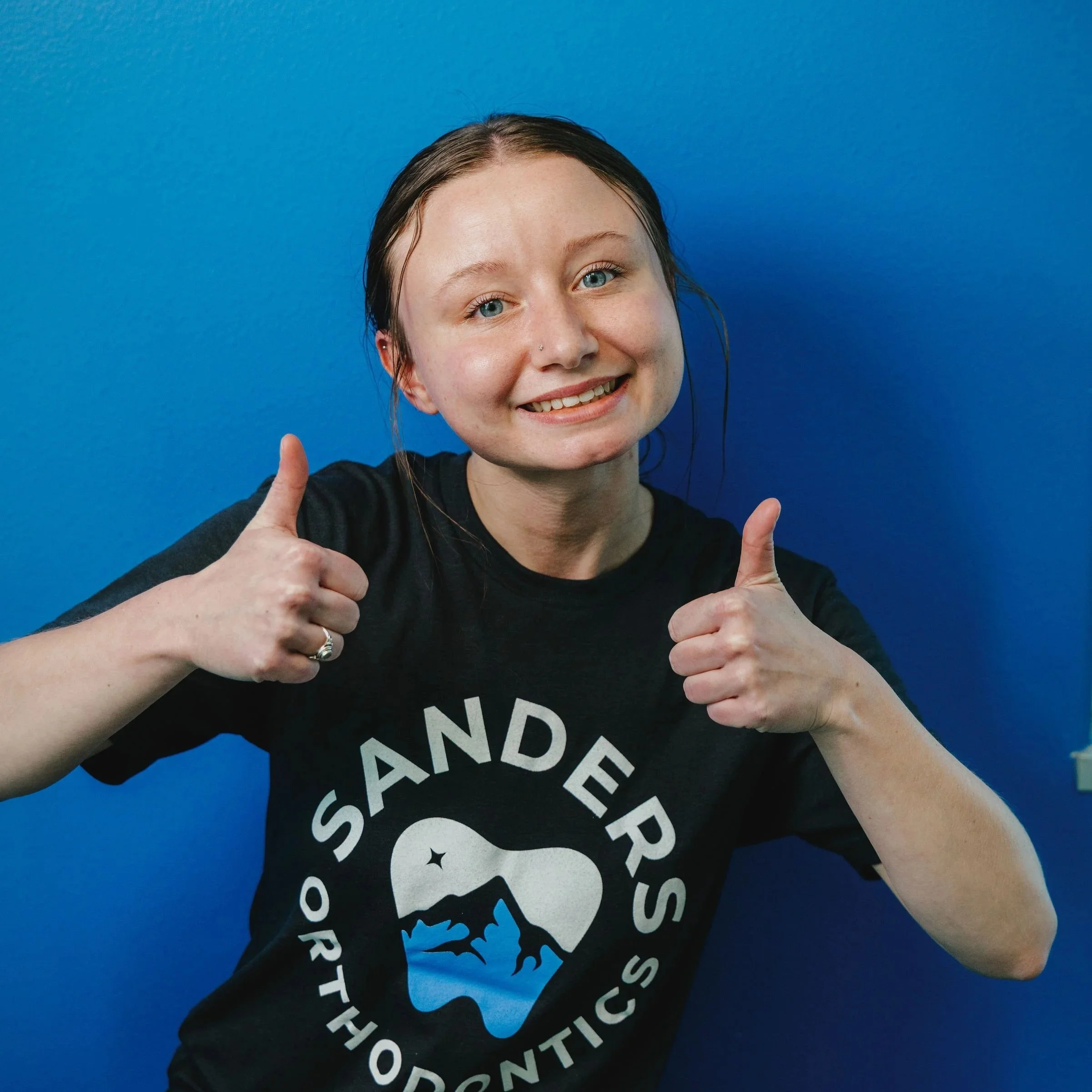 Close-up of a smiling woman with blue eyes and dark hair against a blue background, giving two thumbs up.