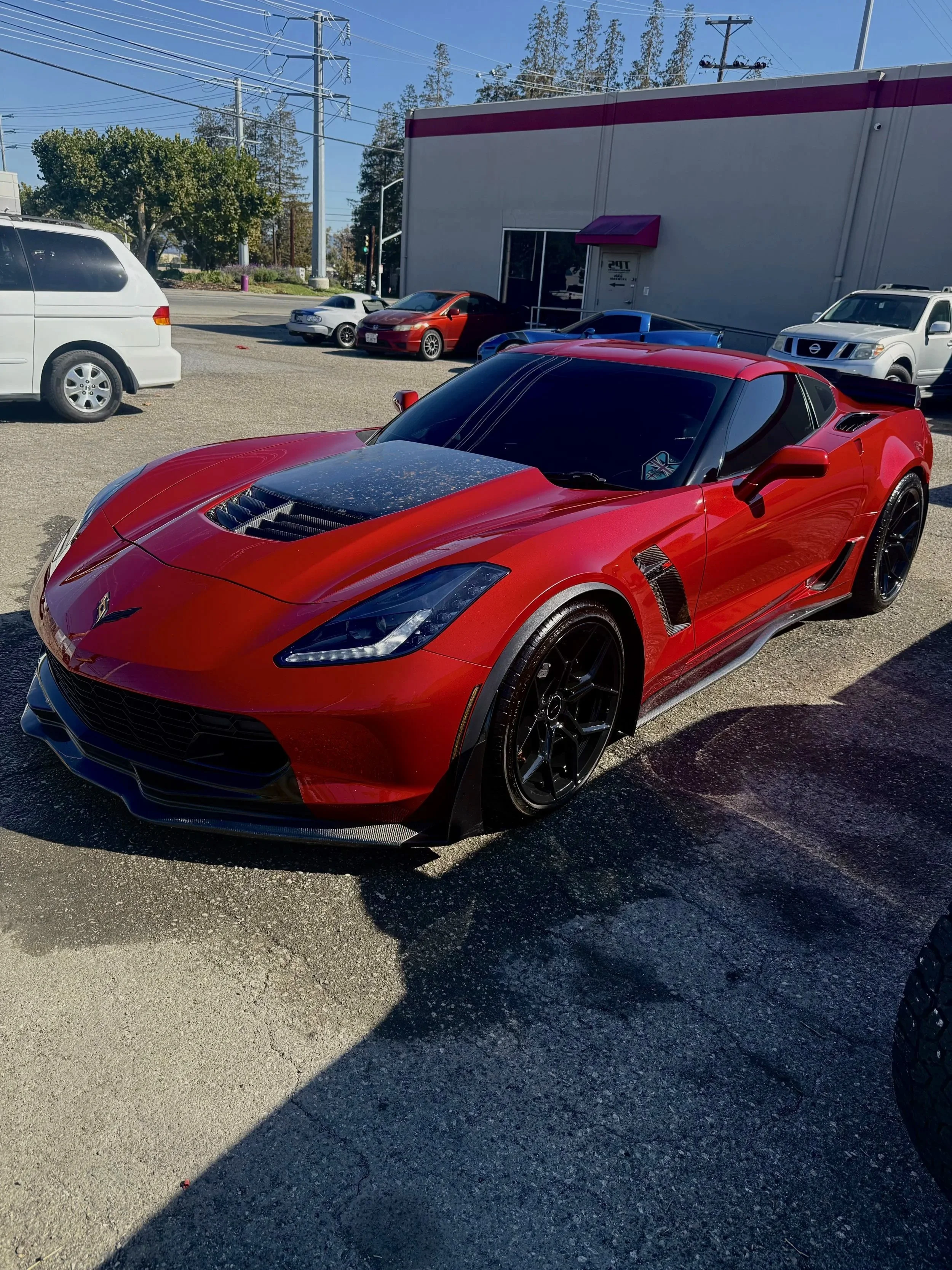 Red sports car with black wheels and carbon fiber accents parked in a lot in front of a commercial building, with other cars and trees in the background.