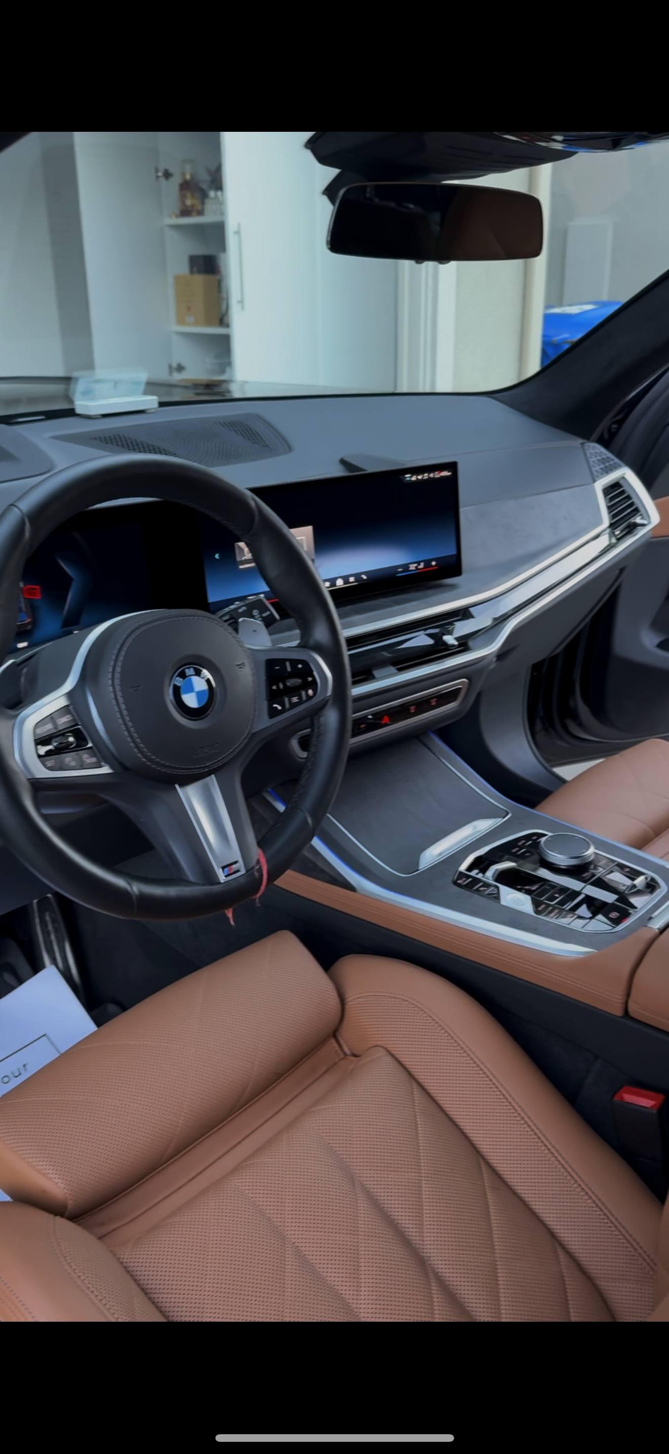 Close-up of the interior of a BMW vehicle, featuring a black steering wheel with the BMW logo, a digital dashboard, an entertainment screen, and tan leather seats.