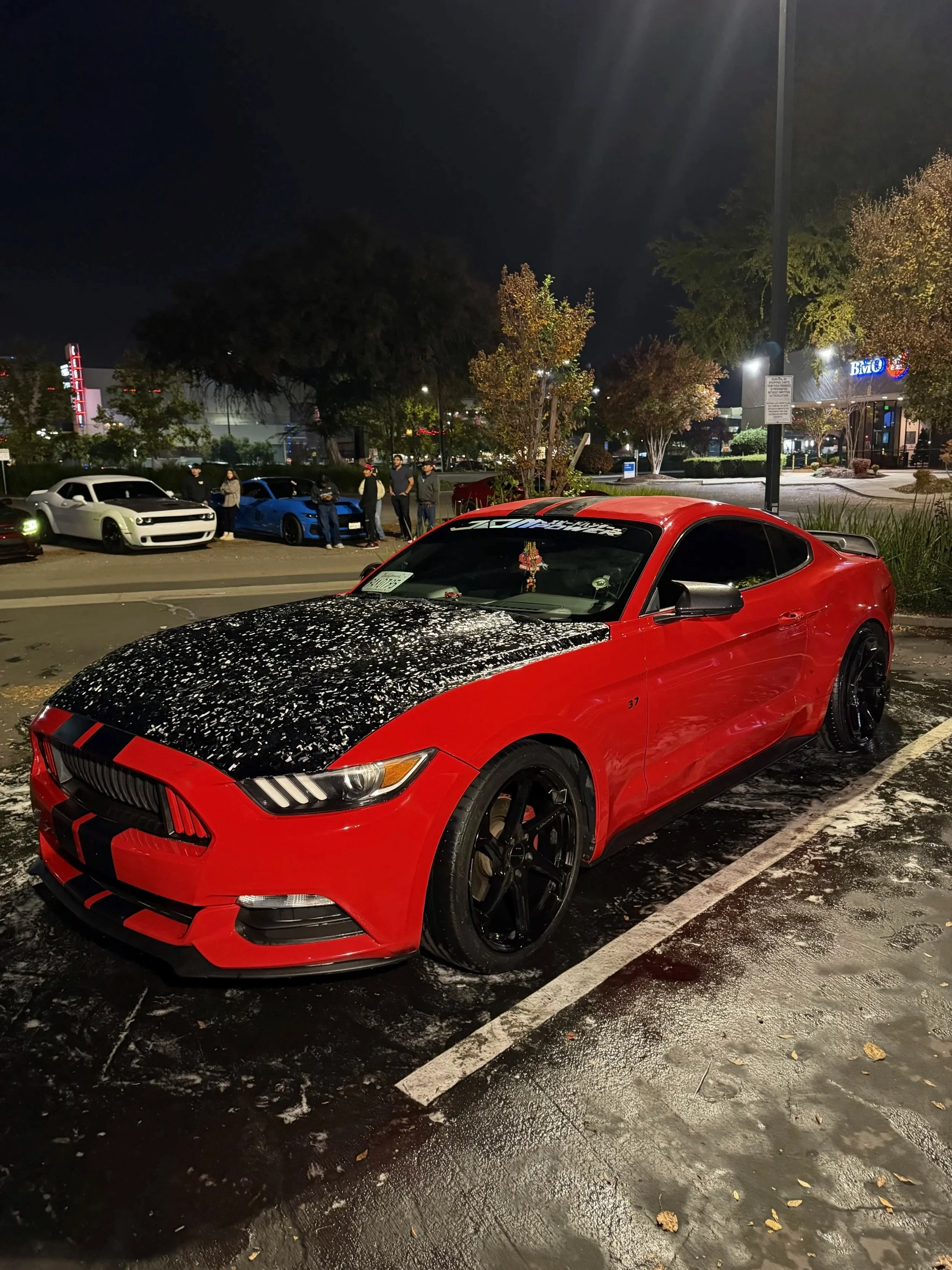 A red sports car with a black hood and black wheels parked at night in a parking lot, with cars and people in the background.