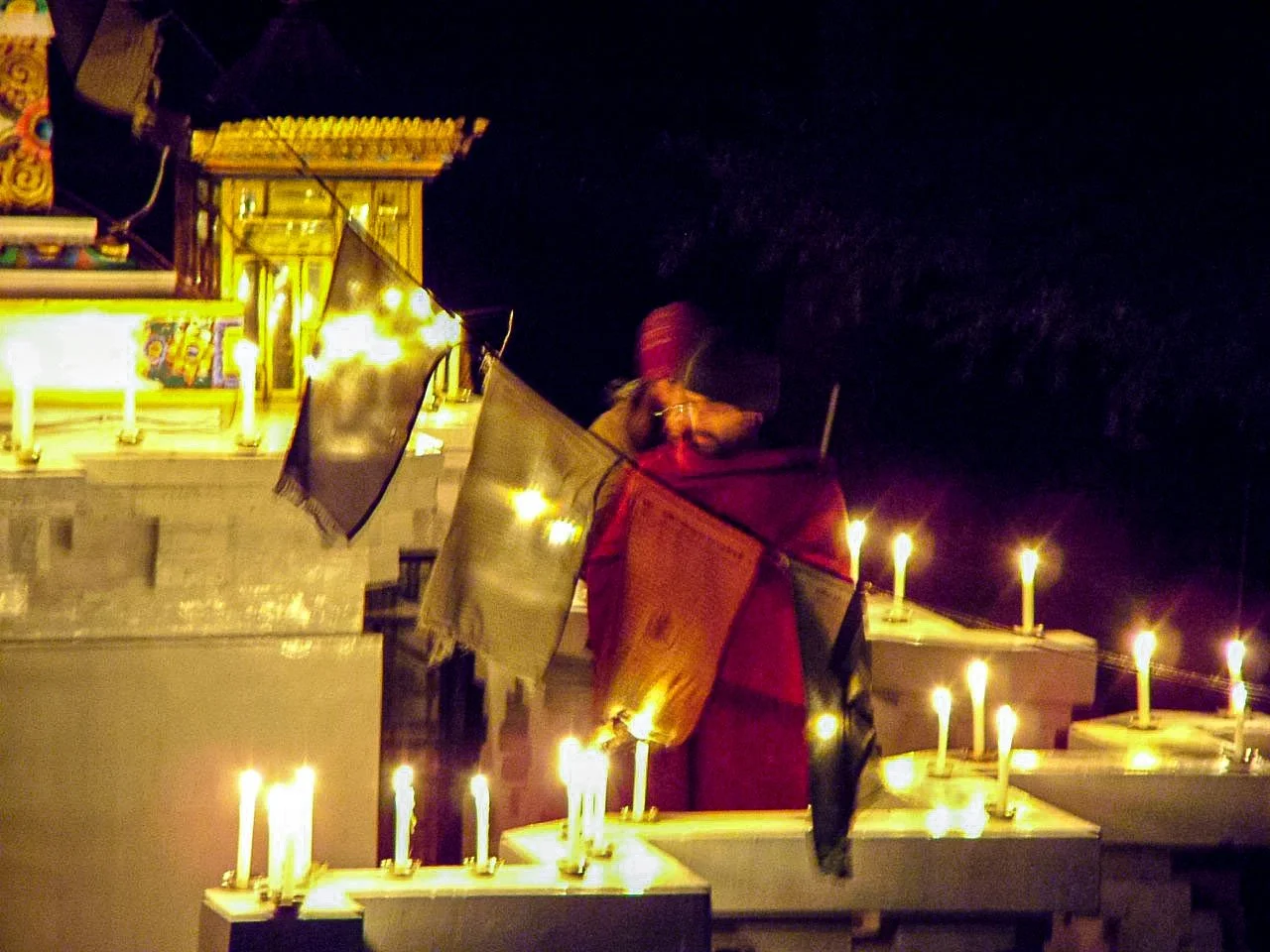 A person dressed in traditional clothing, including a red shawl, lighting candles in a marketplace or temple setting at night. The scene is illuminated by many small candles and ambient lighting.