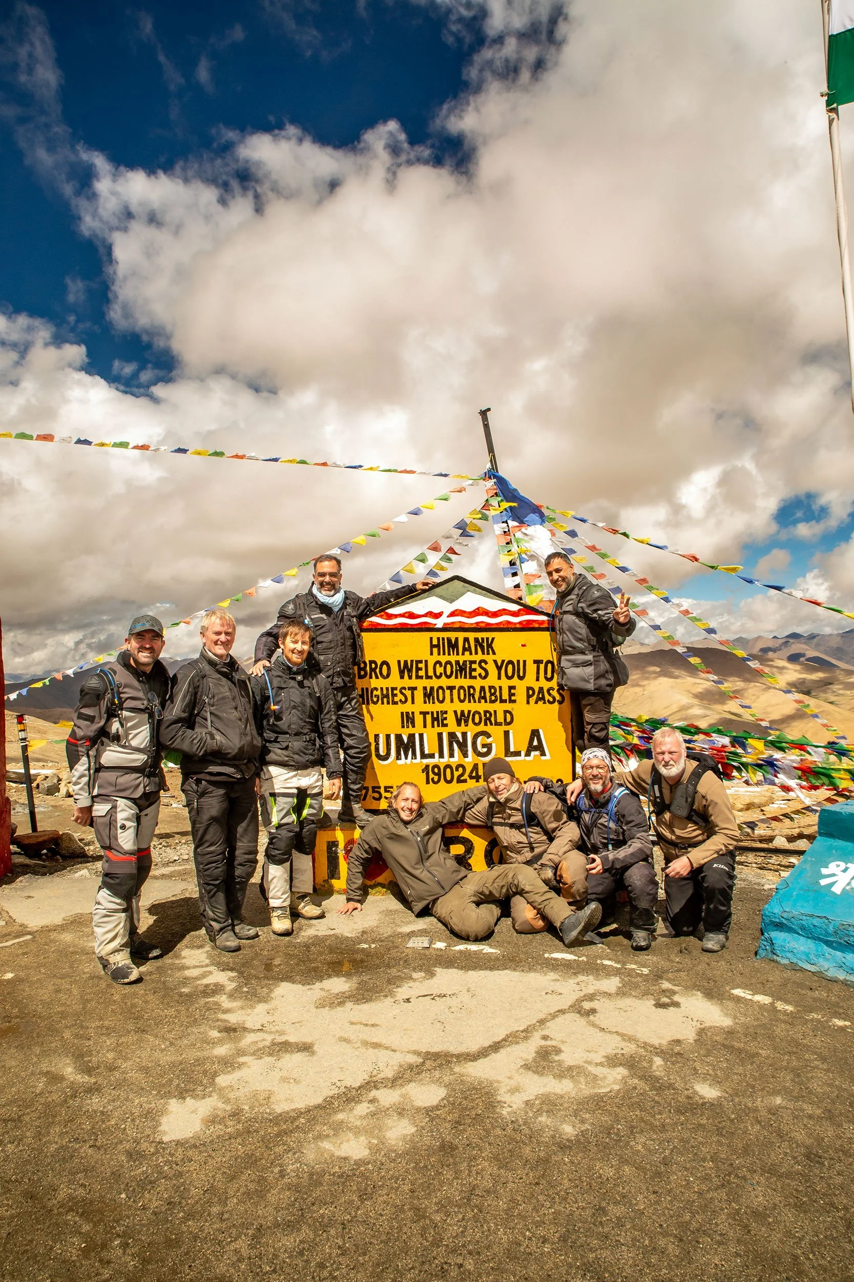 A group of climbers and hikers posing in front of a yellow sign that reads "HIMANK BRO WELCOMES YOU TO HIGHEST MOTORABLE PASS IN THE WORLD. LMULING LA 19024" with colorful prayer flags above them. The sky is partly cloudy and the ground is rocky and barren.
