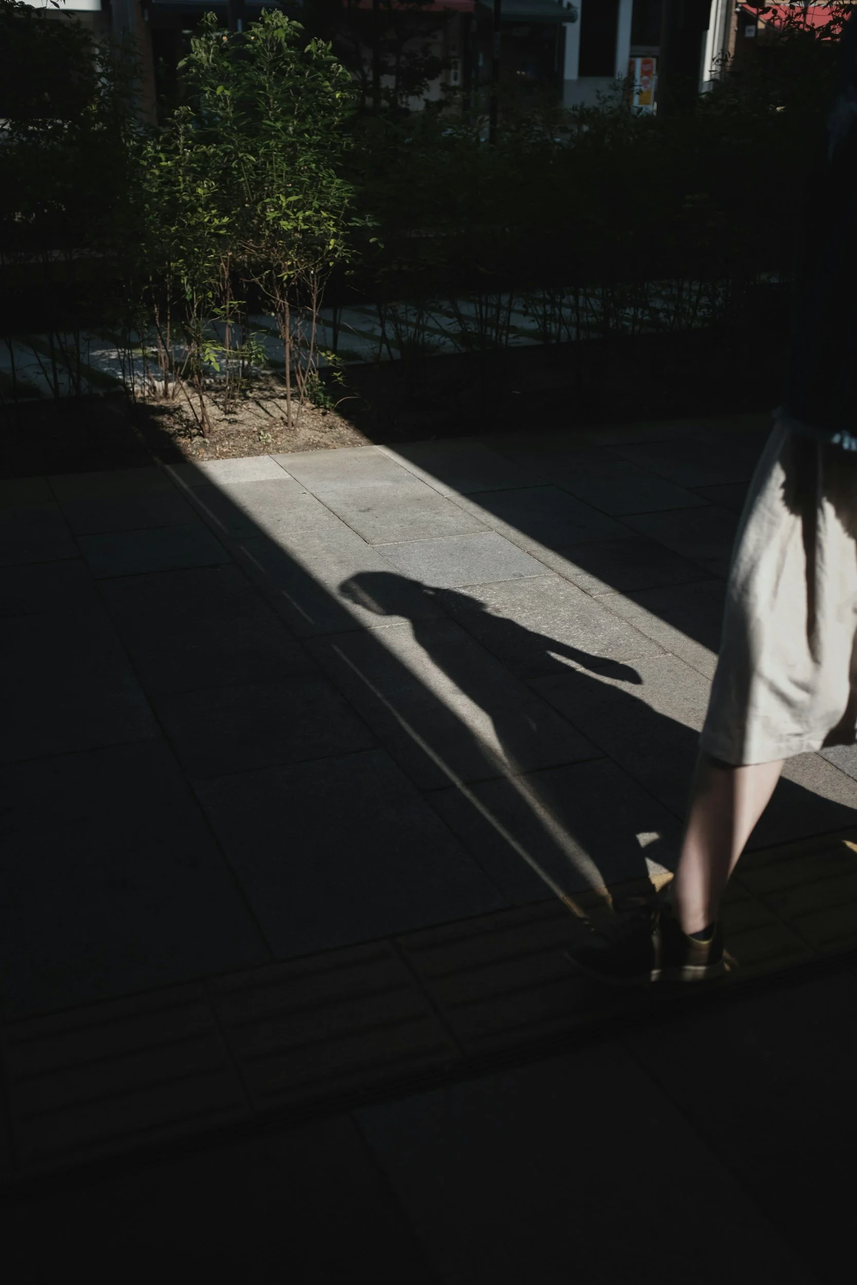 A person wearing shorts and sneakers standing on a shaded outdoor patio with small bushes and a sidewalk in the background.