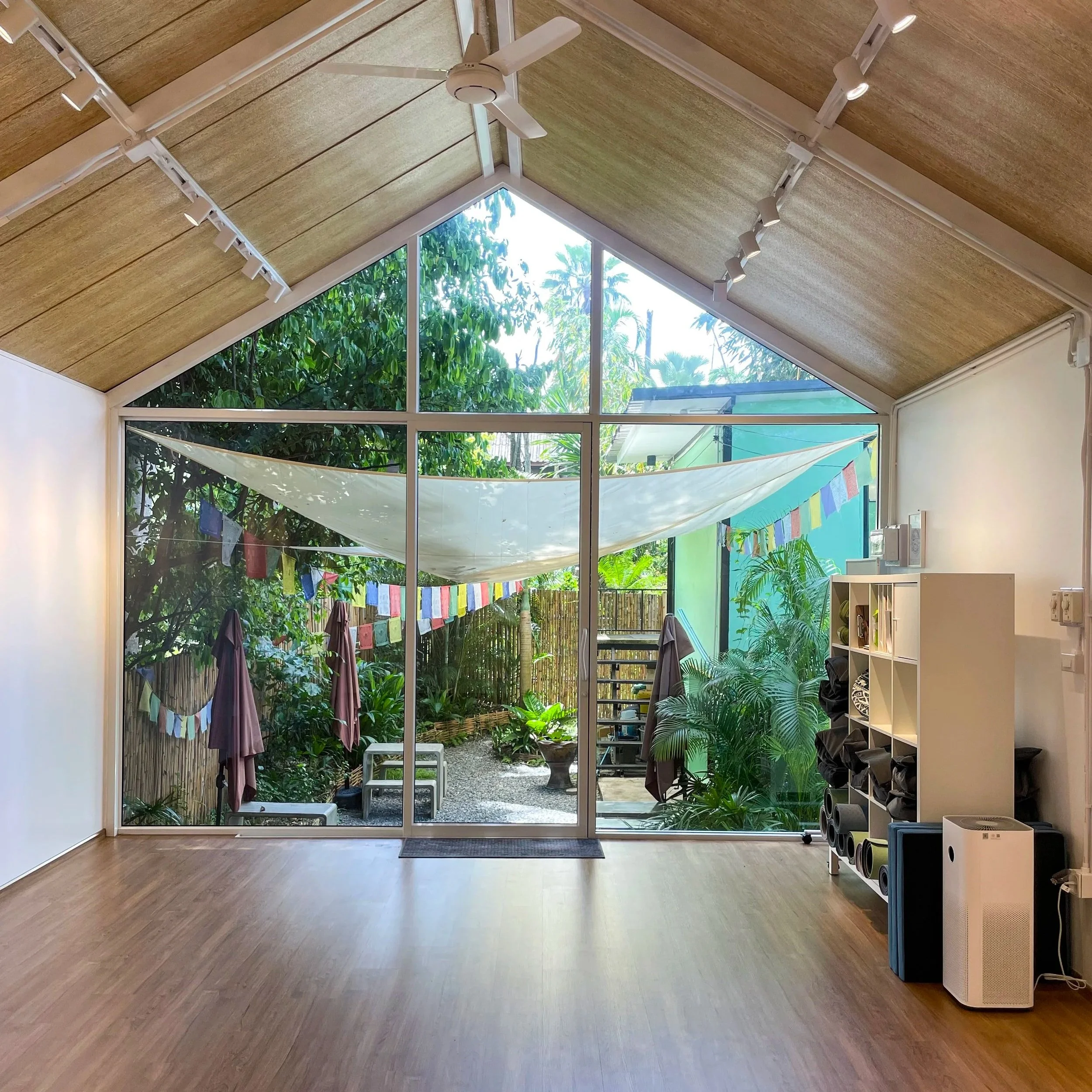 The image shows a room with a garden view through large glass windows, a wooden floor, a white shelving unit with exercise mats, a fan on the ceiling, and colorful flags hanging outside.