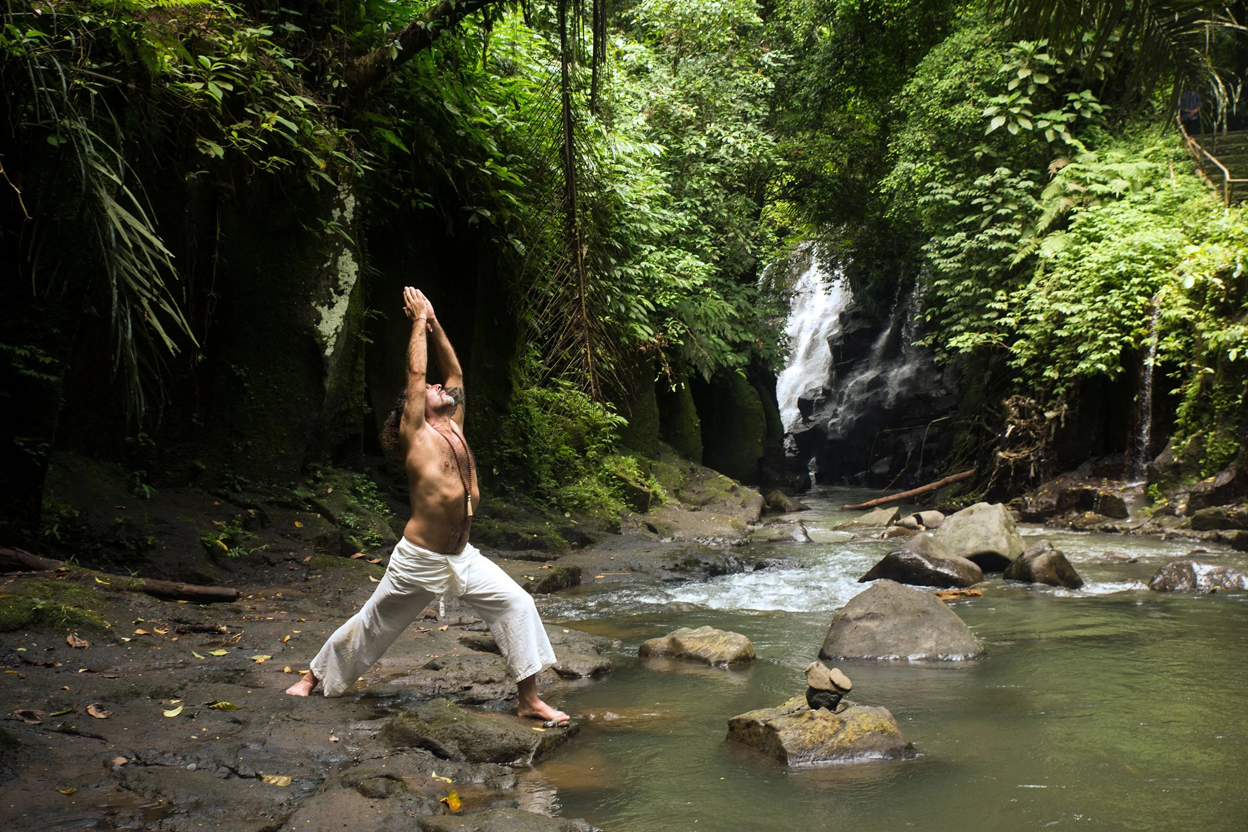 A person with long hair practicing yoga outdoors near a small waterfall in a lush jungle, wearing white pants and shirtless, standing on rocks in a river.