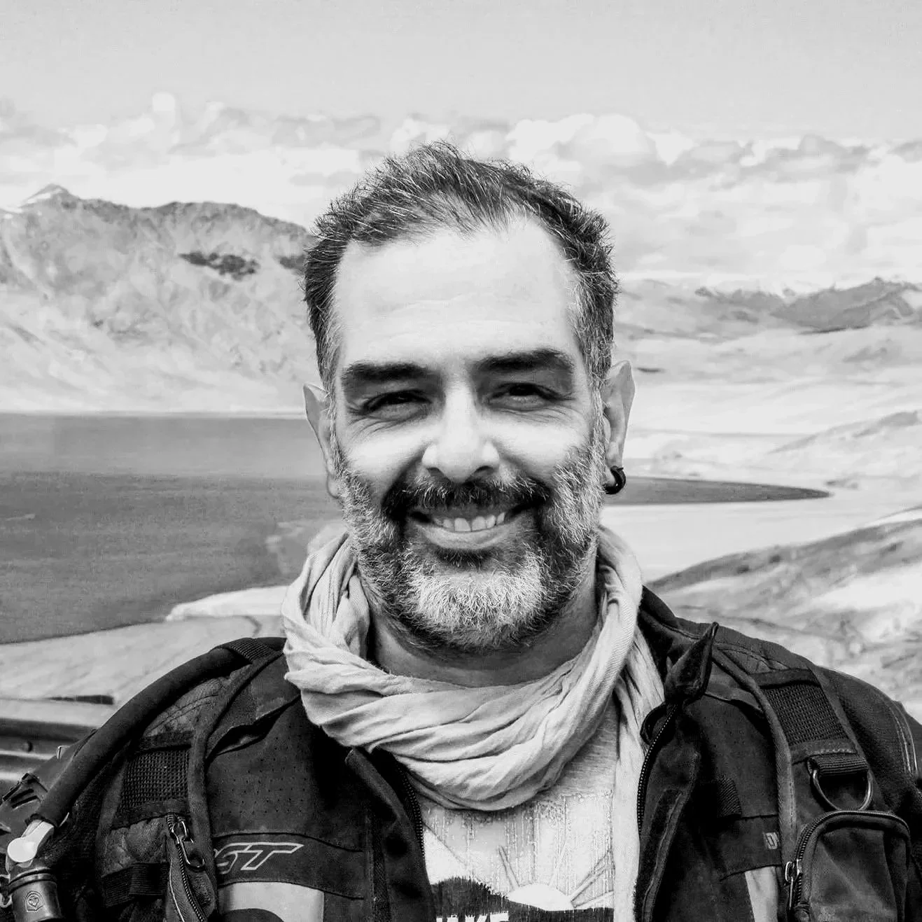 Black and white photo of a smiling man with a beard, short hair, and earring, standing outdoors with mountains and a lake in the background.