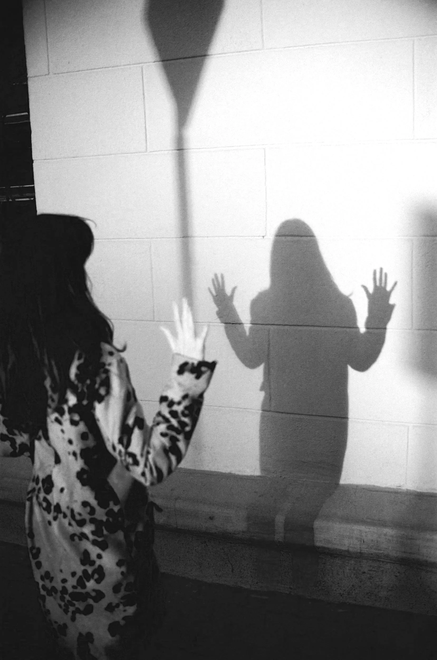 Child with long hair in a cow print outfit touching a wall, casting a shadow of a person with raised hands on the wall.