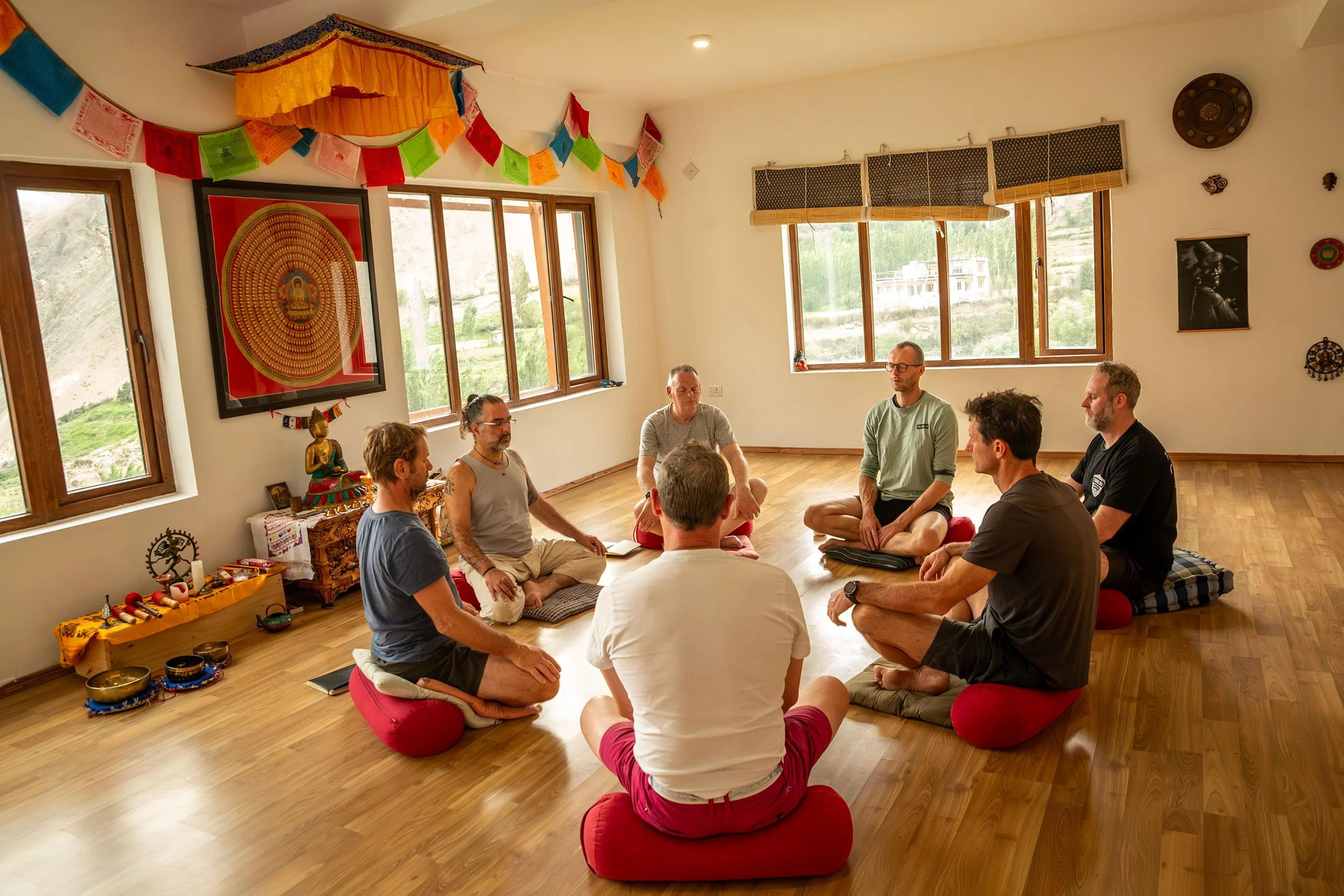 A group of people sitting cross-legged on red cushions in a circle inside a bright room with large windows, spiritual artwork, and decorations on the walls.