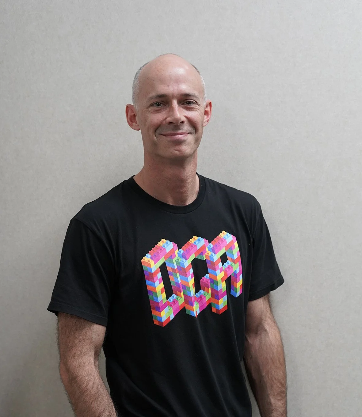 A man with a smile, bald head, wearing a black t-shirt with a colorful 3D lego block design.
