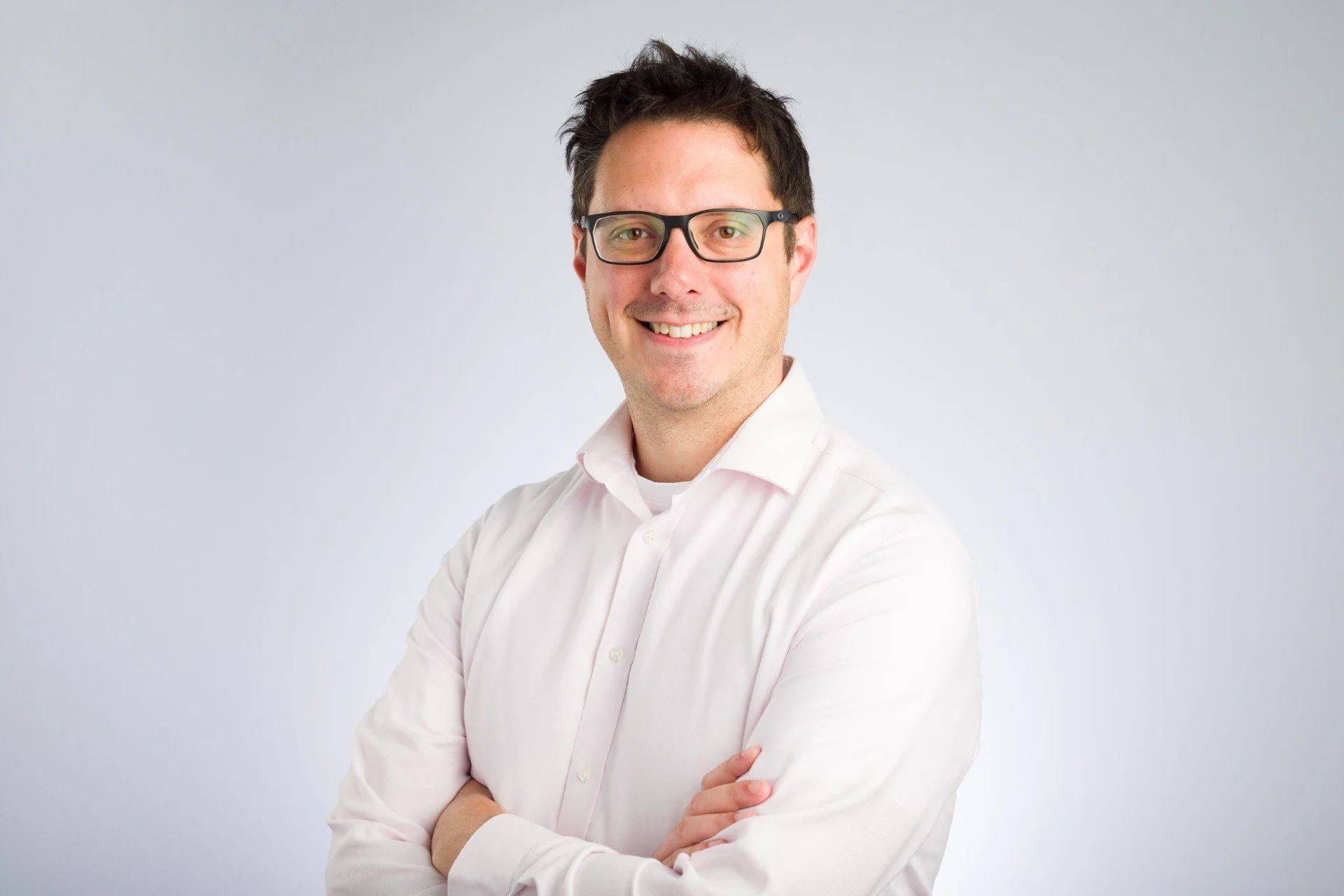 Portrait of a smiling man with dark hair and black glasses, wearing a light pink dress shirt with arms crossed, standing against a plain light gray background.