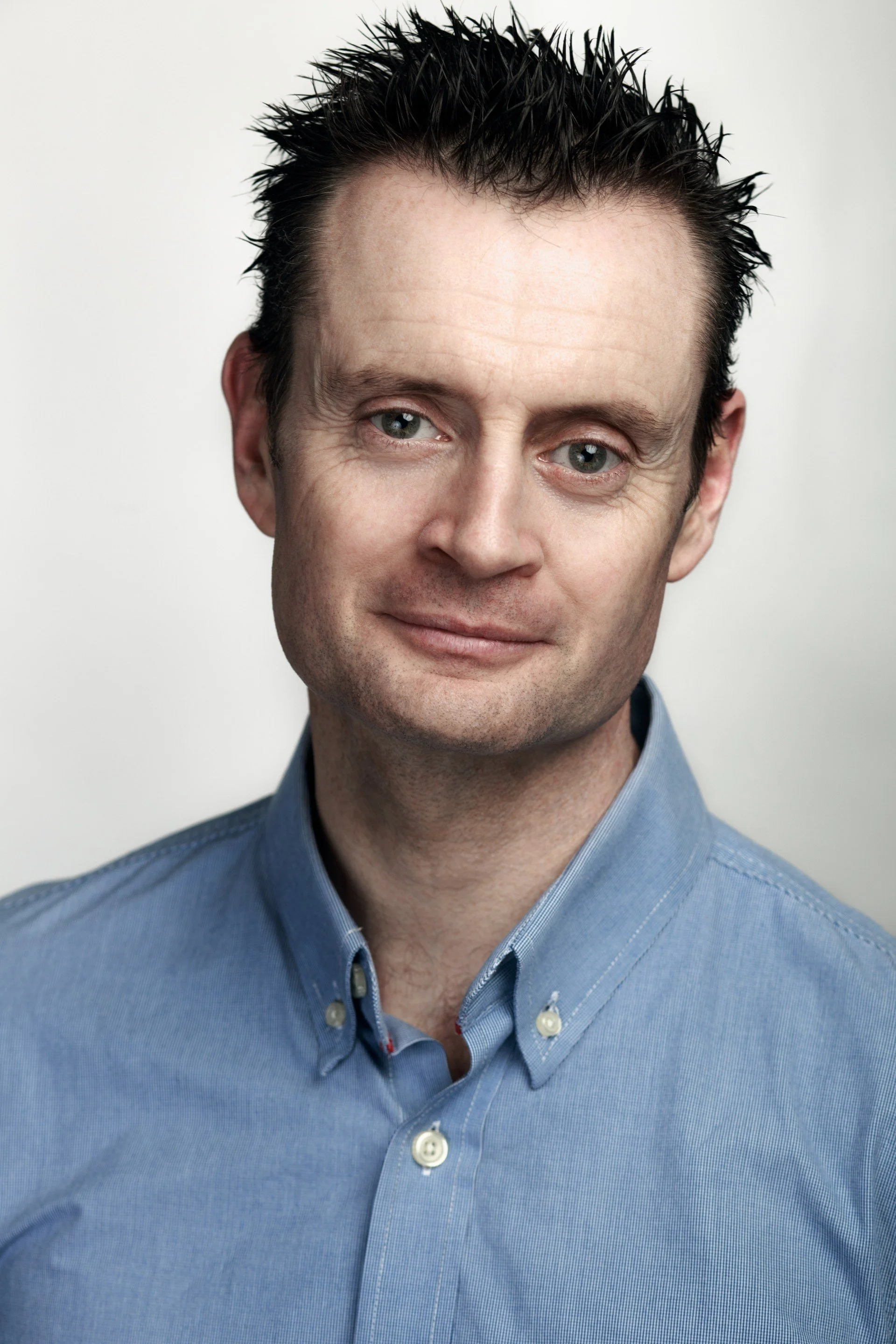 A middle-aged man with short, slightly spiked dark hair, wearing a blue button-up shirt with a collar, smiling gently against a neutral background.