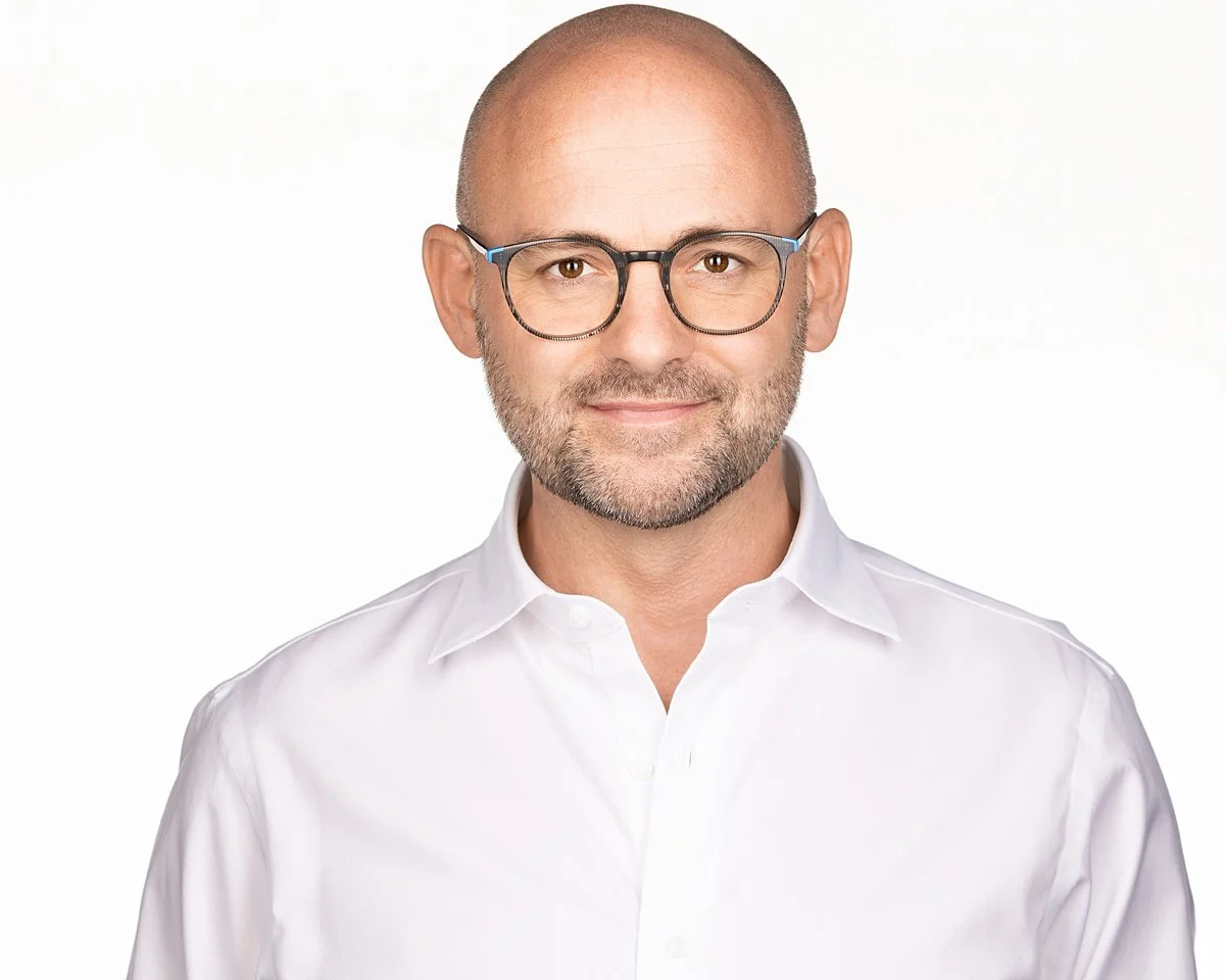 A man with glasses, a beard, and a bald head wearing a white collared shirt, smiling against a plain light background.