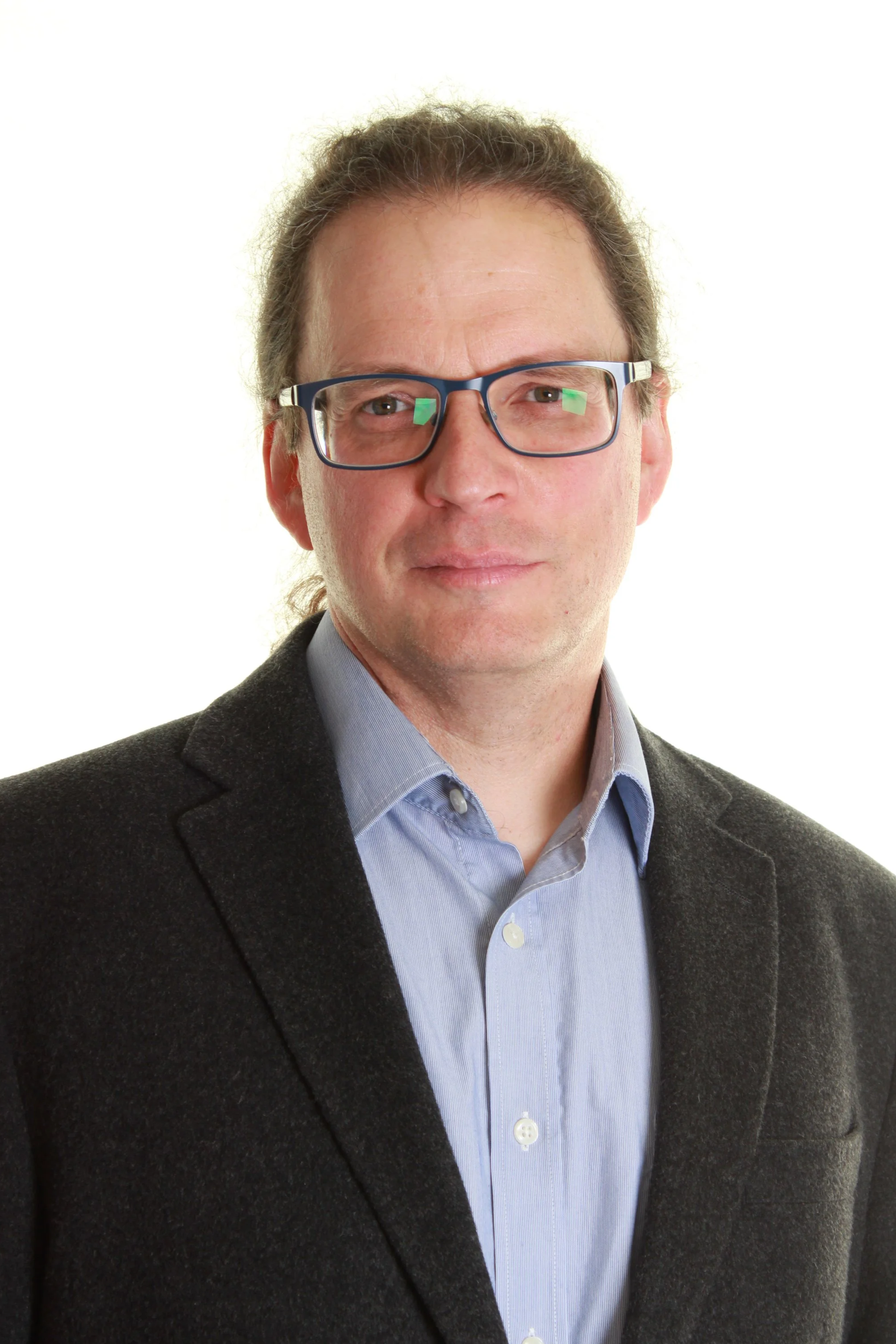A man with glasses wearing a dark blazer over a light blue collared shirt, facing the camera against a white background.