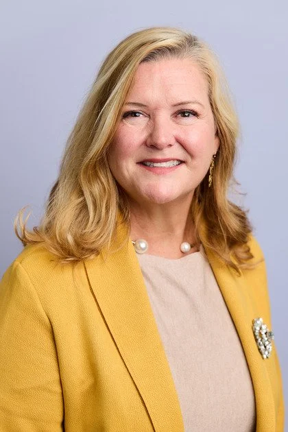 Portrait of a smiling woman with blonde hair wearing a yellow blazer, pearl necklace, and earrings, against a light purple background.