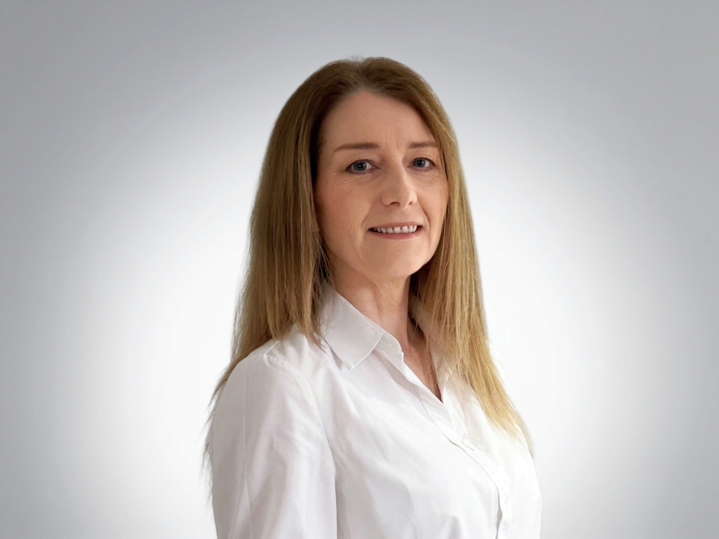A woman with long wavy red hair wearing a white shirt, smiling against a plain light gray background.
