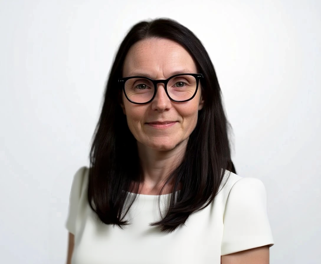 A woman with long dark hair, wearing glasses and a white top, smiling against a plain white background.