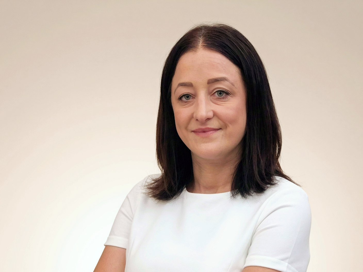 A woman with shoulder-length black hair, light skin, and blue eyes, wearing a white top, standing against a plain light-colored background.