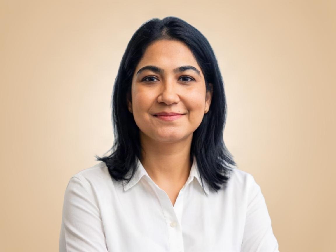 A woman with shoulder-length black hair, wearing a white shirt, smiling with a neutral beige background.
