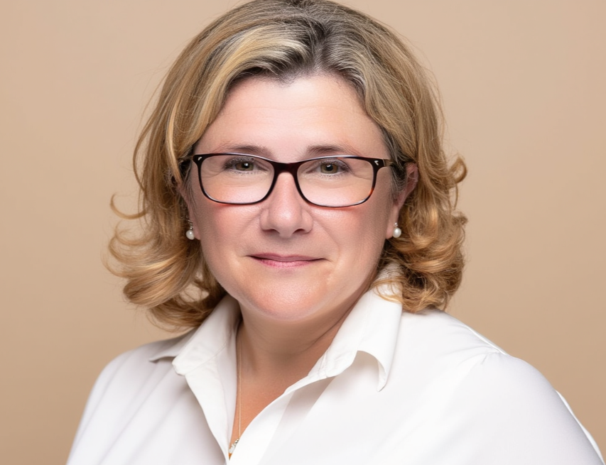 A portrait of a woman with blonde hair, wearing glasses, pearl earrings, and a white shirt, smiling softly against a beige background.
