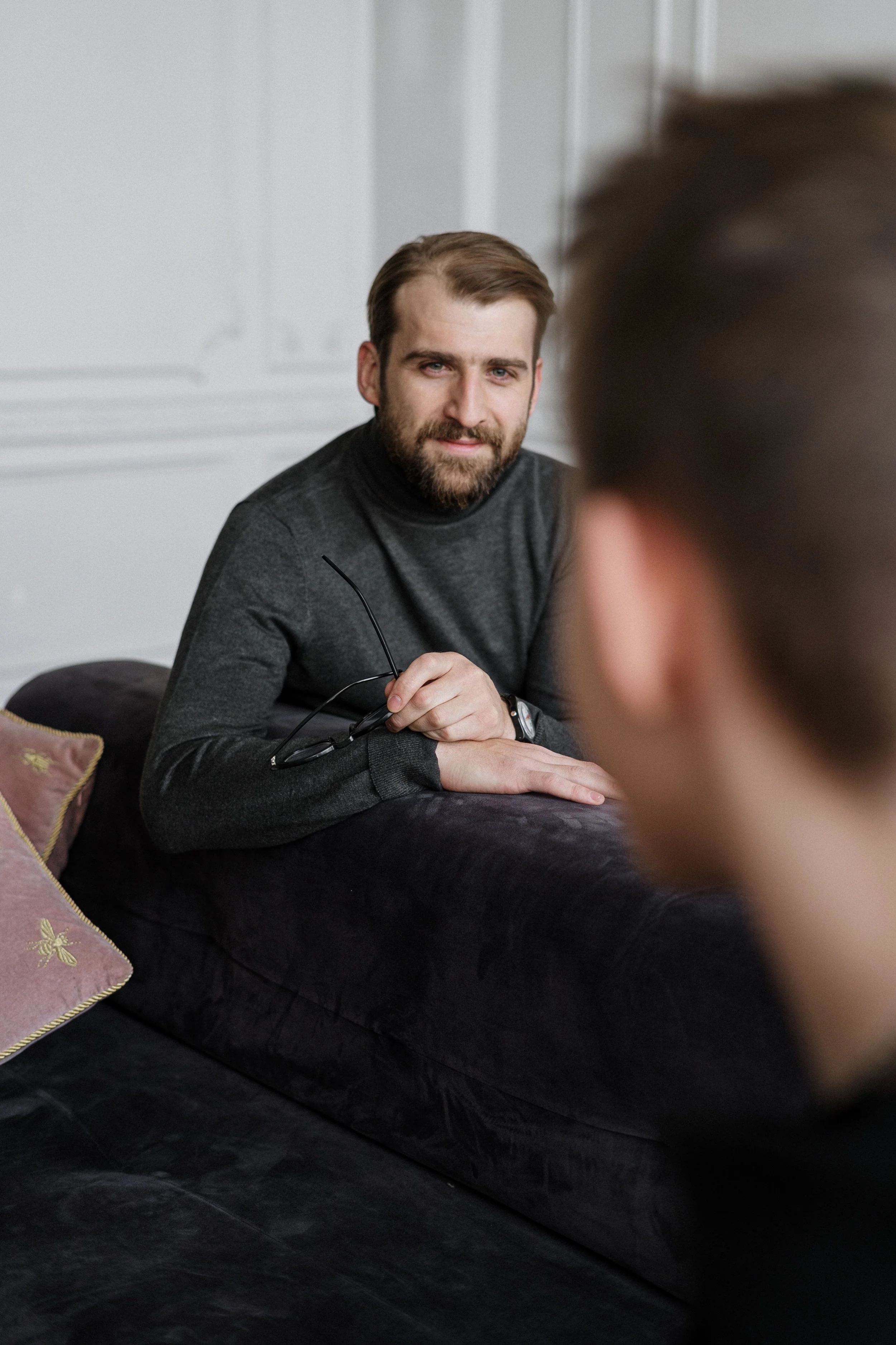 A man with a beard and short hair wearing a dark turtleneck sweater, sitting on a black couch, holding eyeglasses, looking at the camera, with a blurred person in the foreground.